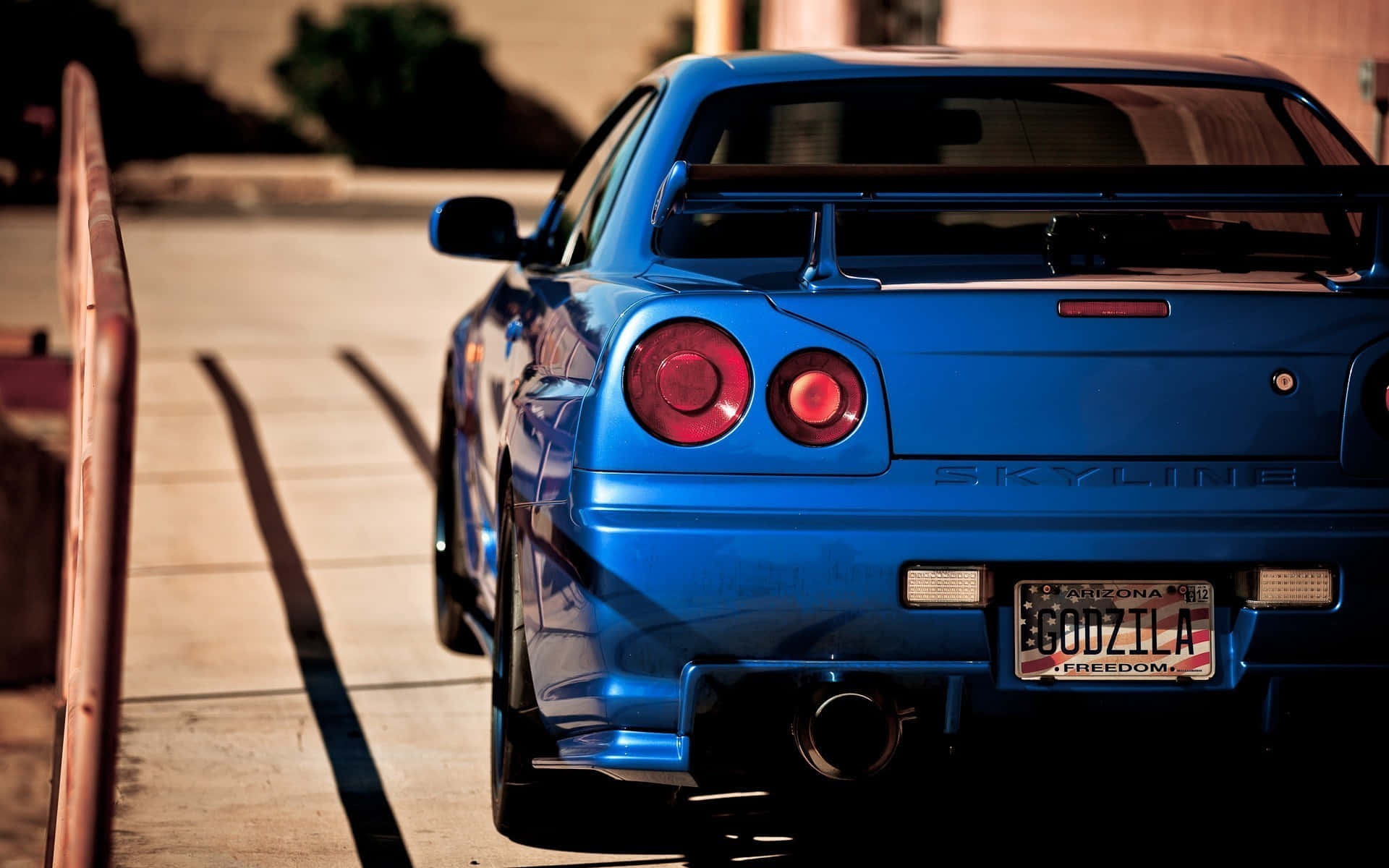 Gtr 桌布 壁紙 — 一輛充滿活力的藍色日產Skyline R34跑車停放在陽光普照的人行道上，帶有Godzilla車牌。