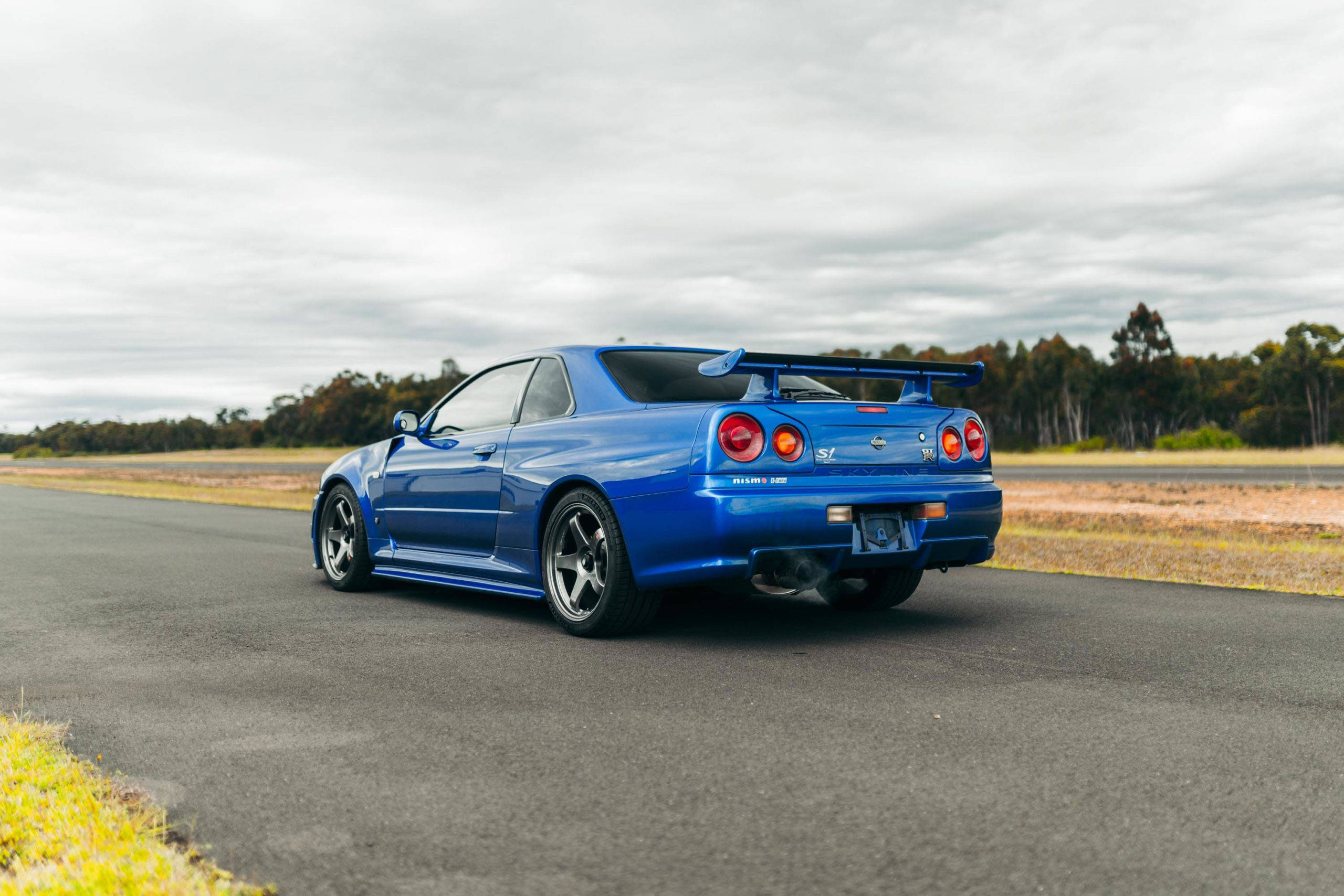 Gtr 桌布 壁紙 — 一輛充滿活力的藍色日產Skyline GT-R R34在陰天下的鄉村柏油路上奔馳。