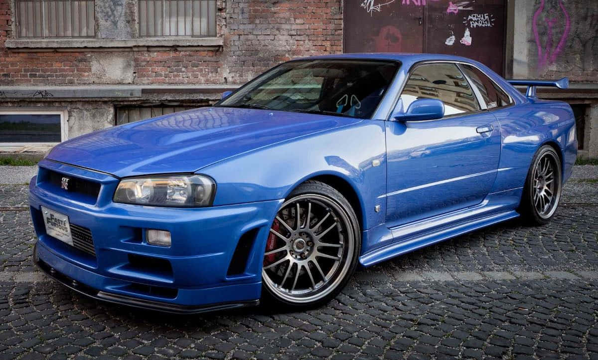 Paul Walker Skyline 桌布 壁紙 — 一輛充滿活力的藍色日產Skyline R34跑車，配備訂製輪圈，停在鵝卵石街道上，旁邊是一棟佈滿塗鴉的磚砌建築。