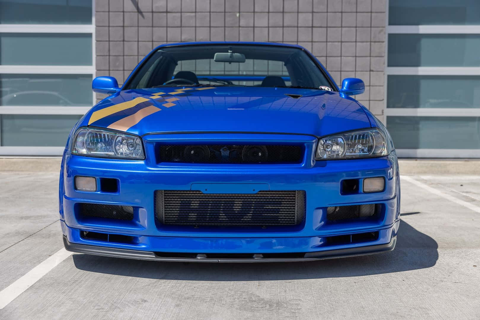 Paul Walker Skyline 桌布 壁紙 — 一輛充滿活力的藍色日產Skyline R34跑車，帶有黃色和黑色賽車條紋，停在一棟現代建築前。