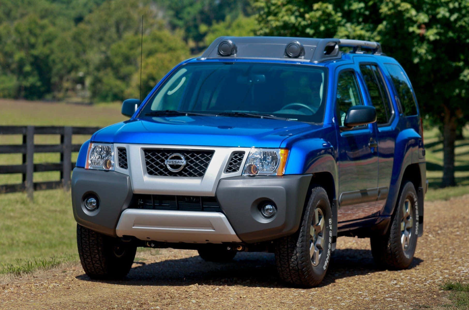 Nissan Xterra 桌布 壁紙 — 一輛充滿活力的藍色日產Xterra SUV，配備車頂行李架，停在陽光明媚的鄉村田野中一條碎石路旁，旁邊是木柵欄。