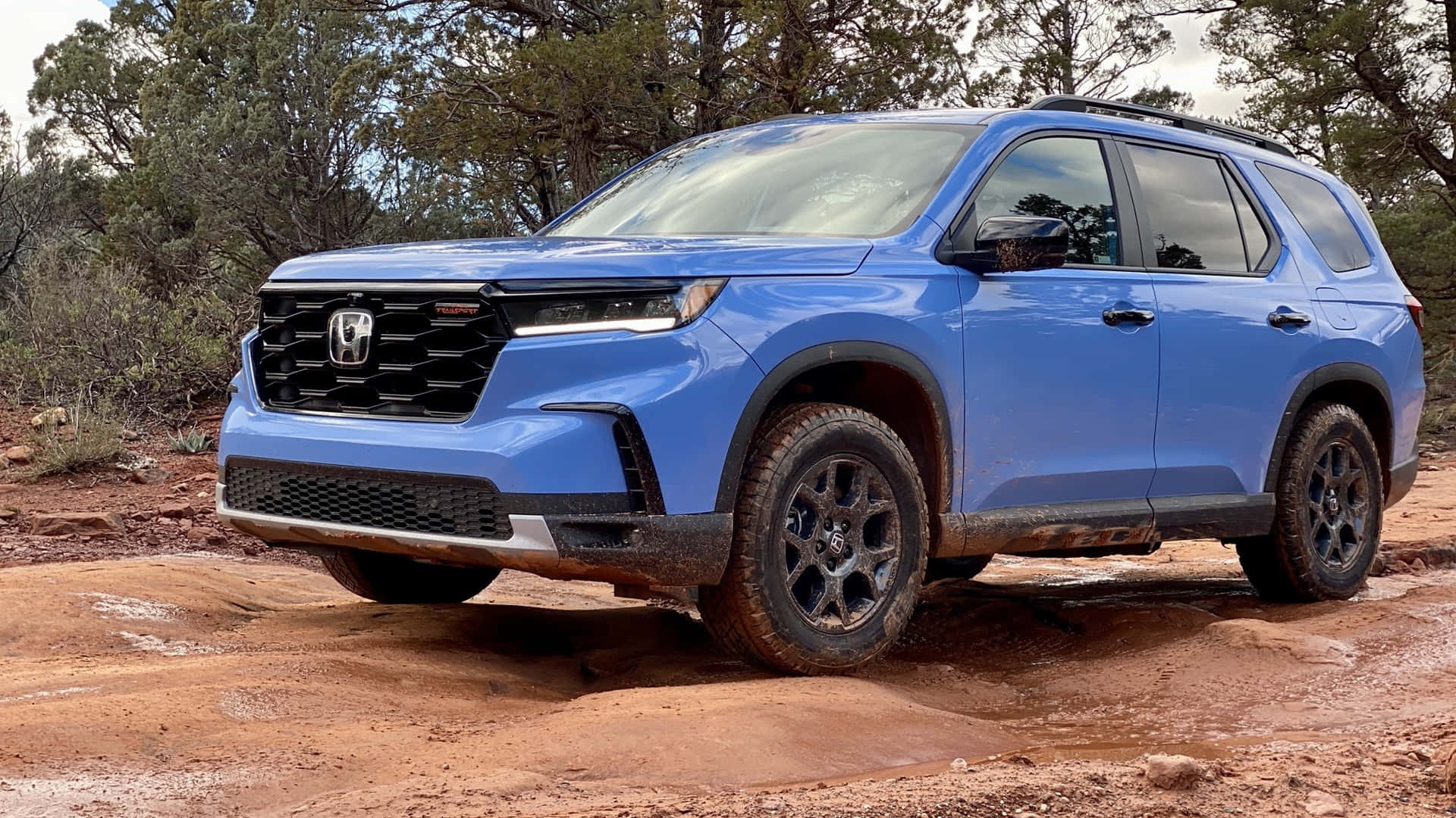 Honda Pilot 桌布 壁紙 — 一輛充滿活力的藍色本田SUV配備黑色輪胎，在多雲的天空下行駛於泥濘的紅色岩石地形。