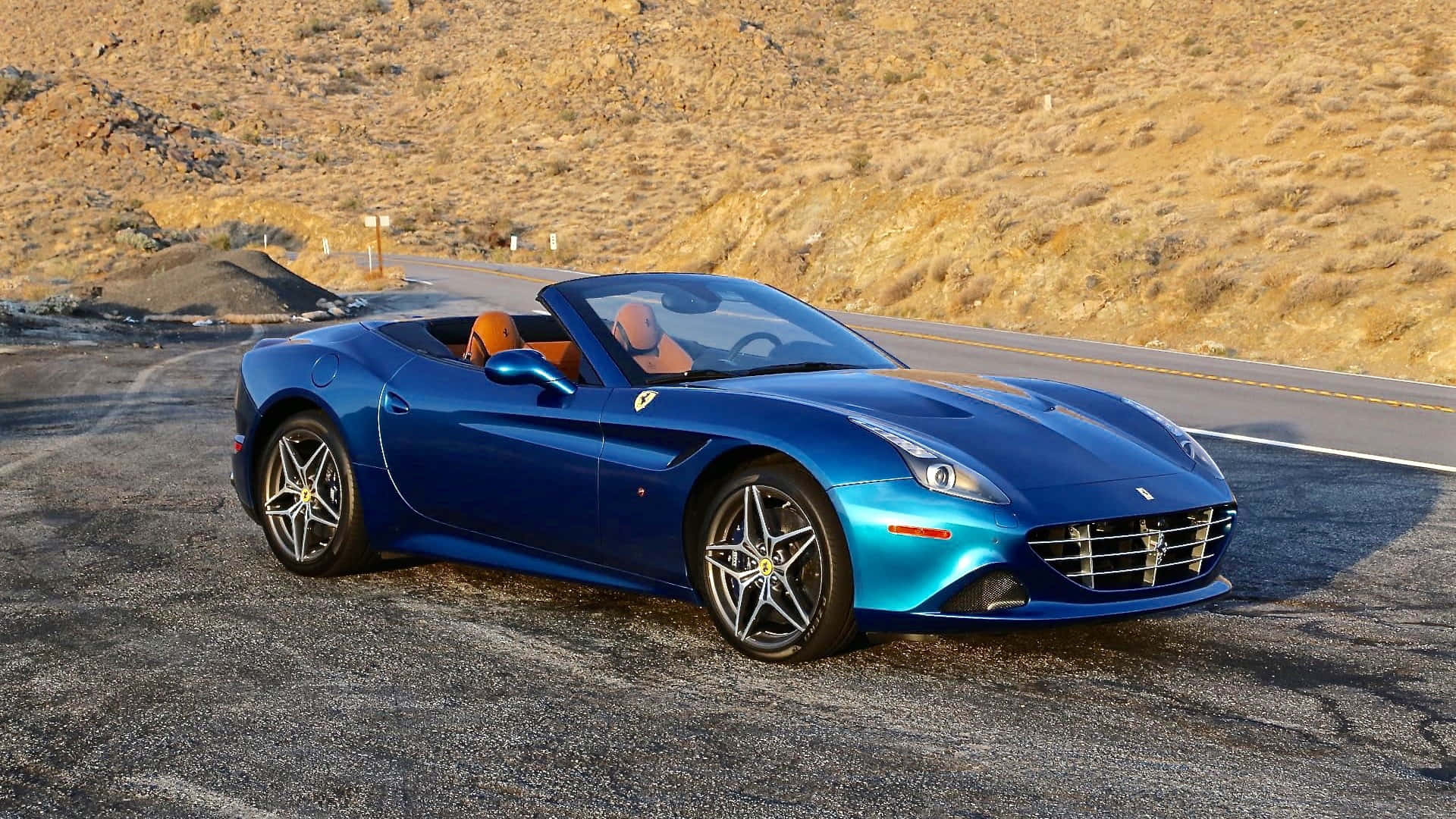 Ferrari California T 桌布 壁紙 — 一輛充滿活力的藍色法拉利敞篷車停在沙漠公路上，背景是金色的山丘。