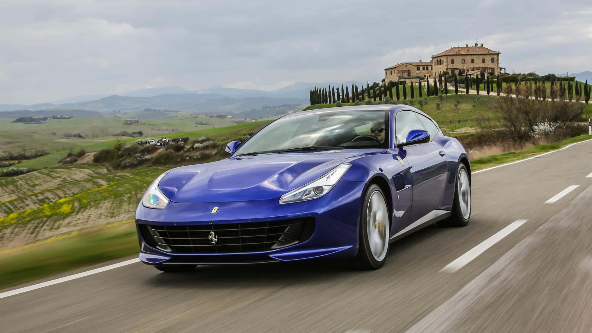 Ferrari Gtc4lusso 桌布 壁紙 — 一輛充滿活力的藍色法拉利跑車沿著蜿蜒的道路穿過起伏的綠色山丘，背景是一座石頭別墅坐落在山頂上。