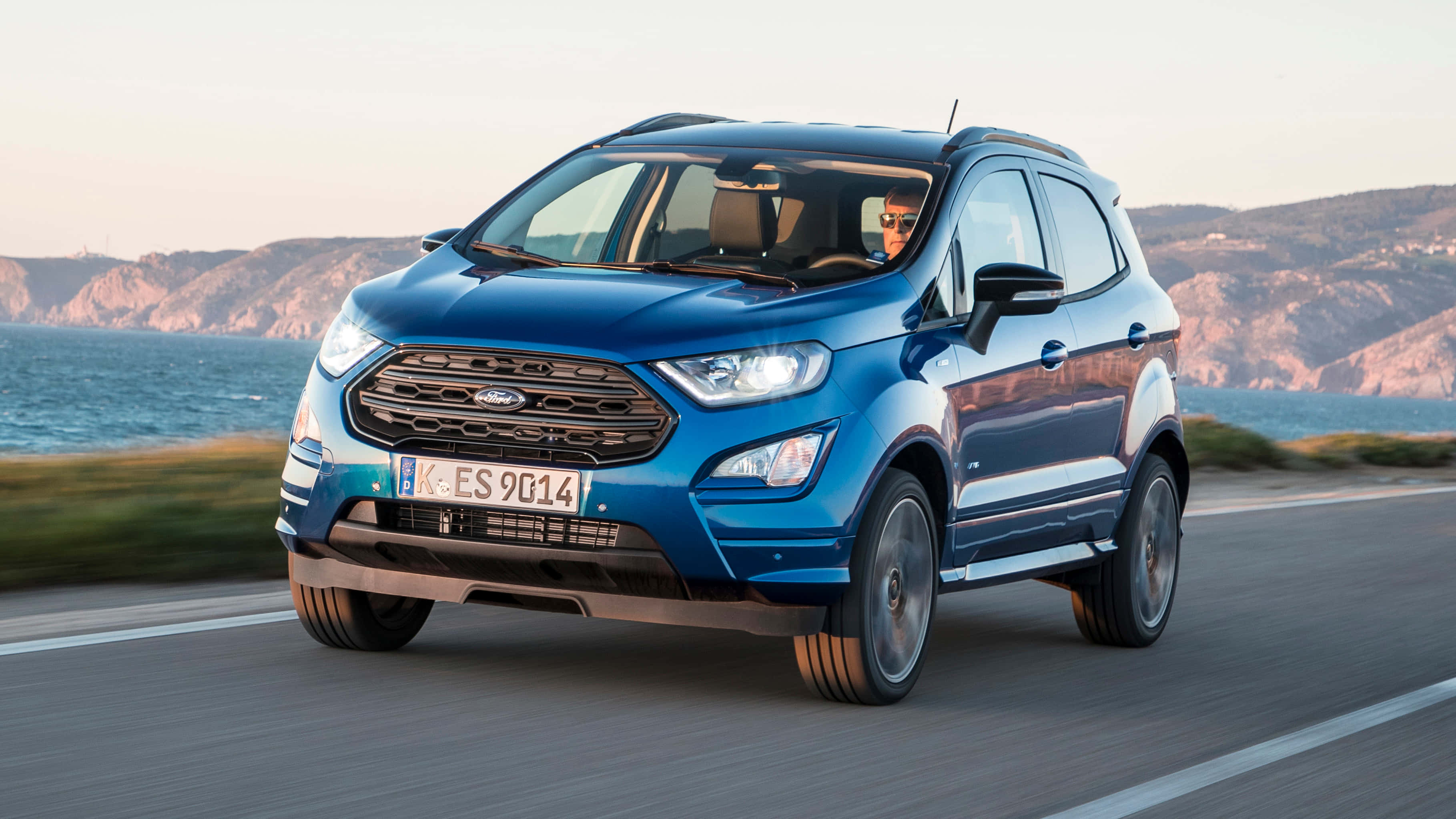 福特 桌布 壁紙 — 一輛充滿活力的藍色福特 EcoSport SUV 沿著海岸道路行駛，背景是海洋和山丘。
