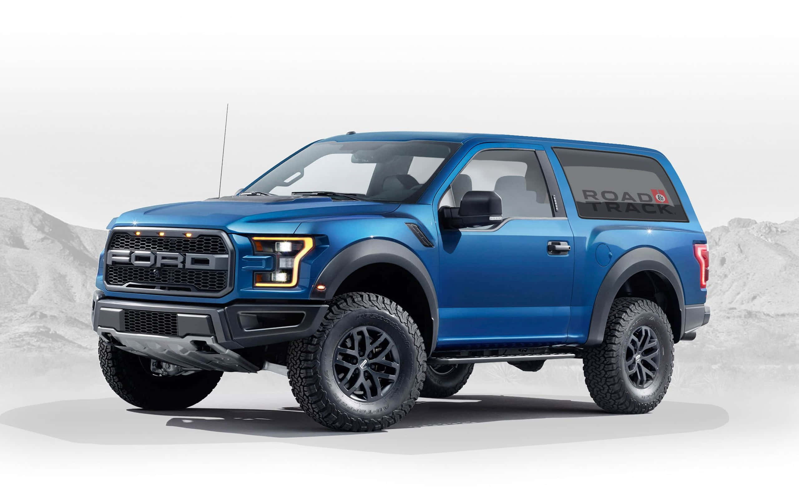 福特野馬越野車 桌布 壁紙 — 一輛充滿活力的藍色福特 F-150 Raptor SUV，帶有黑色飾邊和「ROAD TRACK」貼花，在荒涼的山景背景下蓄勢待發。