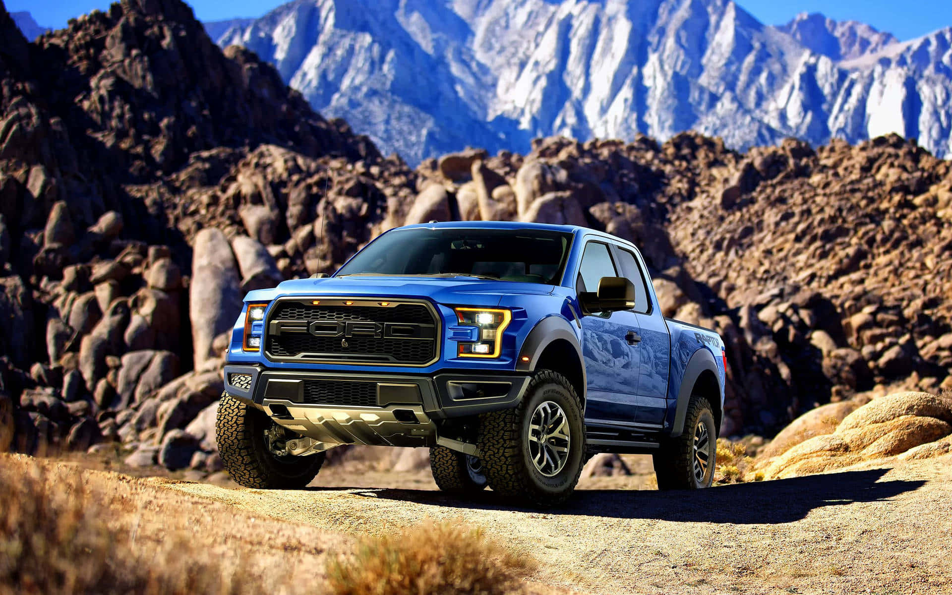 福特 桌布 壁紙 — 一輛充滿活力的藍色福特 F-150 Raptor 卡車，在晴朗的藍天下，停放在崎嶇的沙漠小徑上，岩石地形後方聳立著白雪覆蓋的山峰。