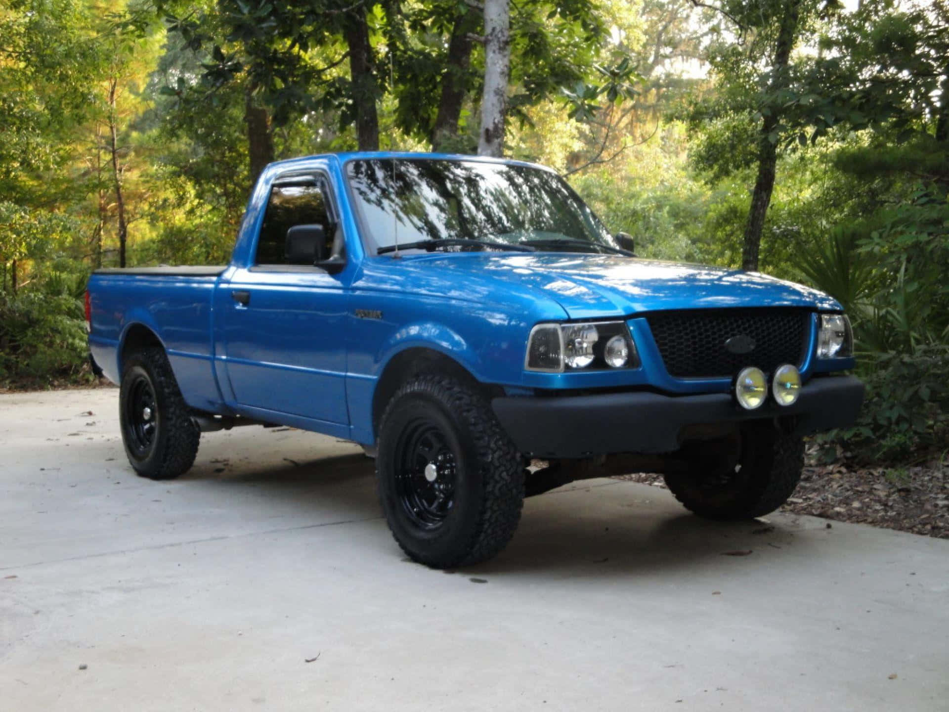 Ford Ranger 桌布 壁紙 — 一輛充滿活力的藍色福特 Ranger 皮卡車，配有黑色車輪和前保險桿燈，停在混凝土車道上，周圍環繞著茂密的綠色樹木。