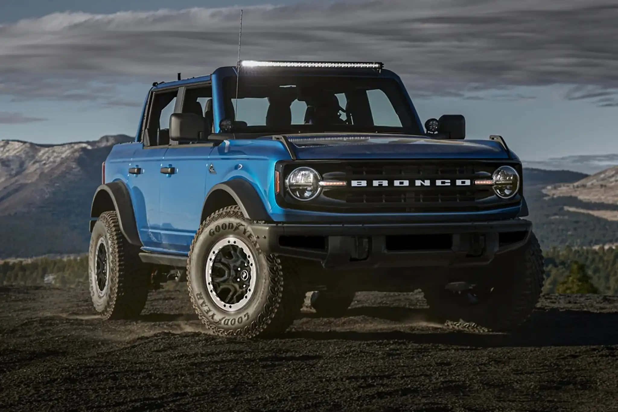 福特野馬越野車 桌布 壁紙 — 一輛充滿活力的藍色福特Bronco SUV在崎嶇地形上大膽地矗立著，背景是白雪覆蓋的山脈和陰雲密布的天空。