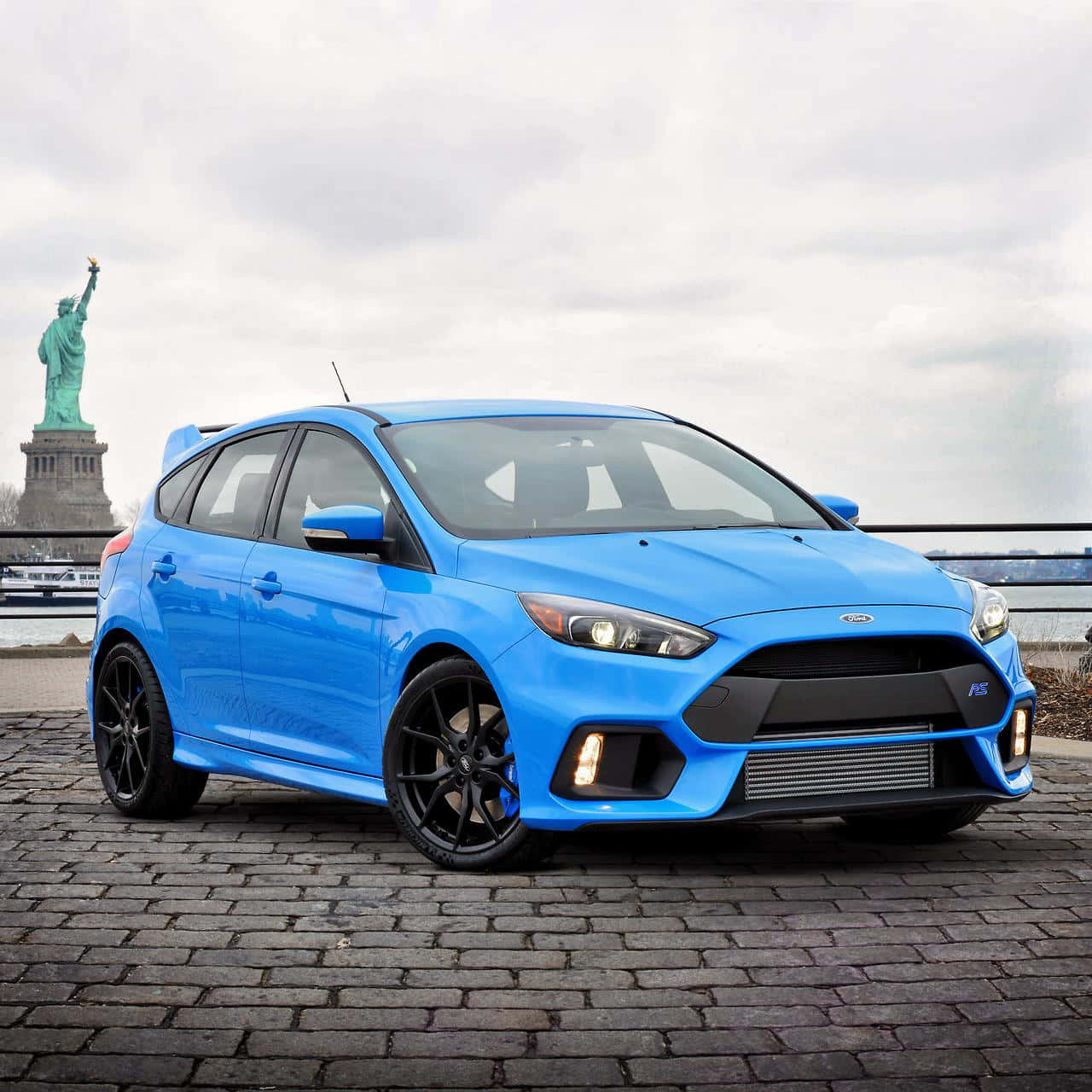 Ford Focus 桌布 壁紙 — 一輛充滿活力的藍色福特福克斯RS停在鵝卵石路上，背景是陰天下的自由女神像。
