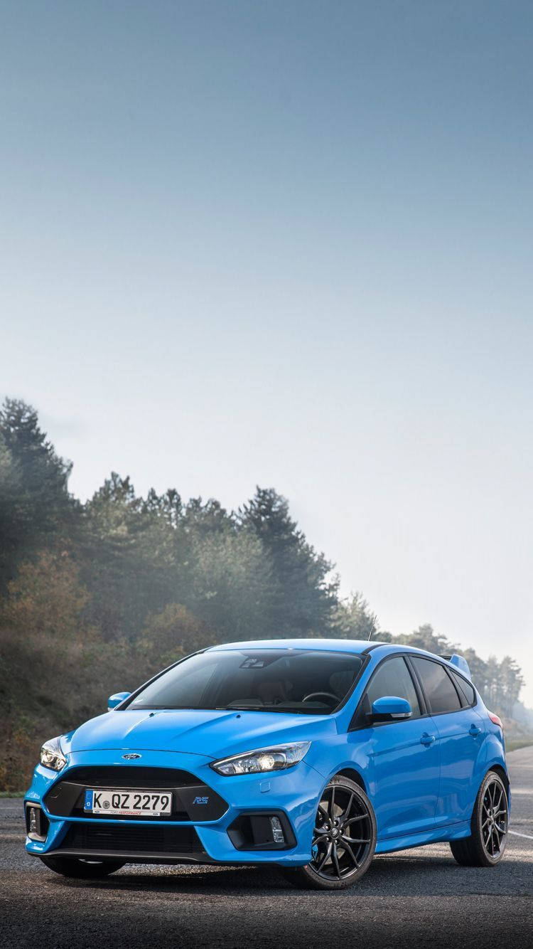 ford iPhone 桌布 壁紙 — 一輛充滿活力的藍色福特福克斯RS跑車停在瀝青路上，周圍是森林覆蓋的山丘，天空晴朗。
