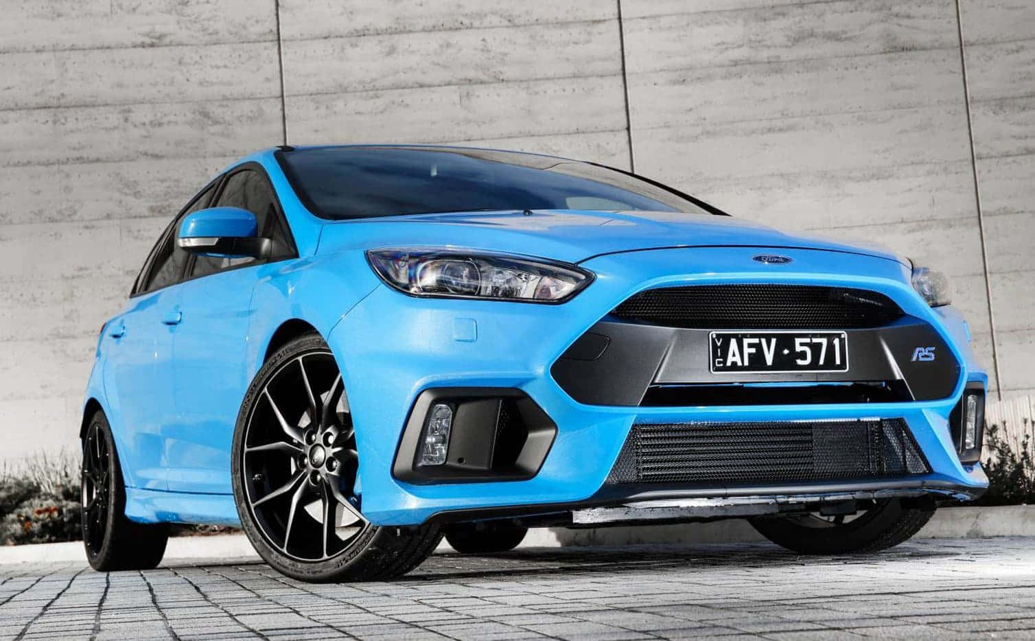 Ford Focus 桌布 壁紙 — 一輛充滿活力的藍色福特福克斯RS跑車，帶有黑色飾邊和定制輪轂，停放在石鋪路面上，背靠混凝土牆。