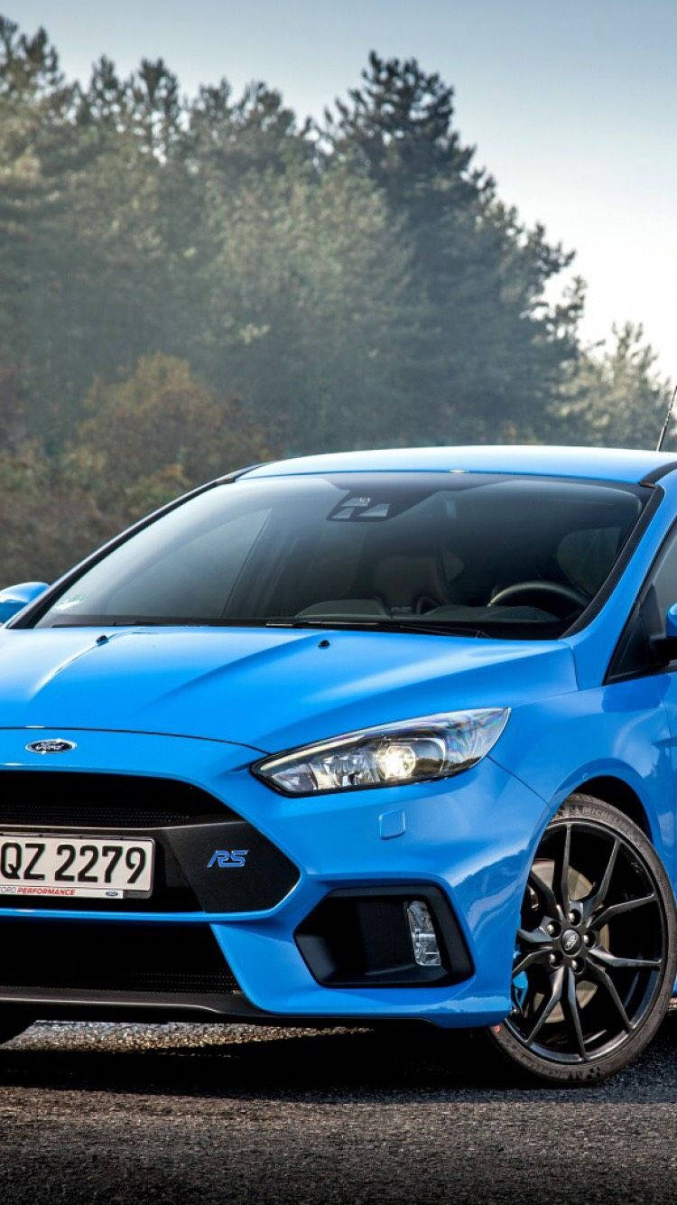 ford iPhone 桌布 壁紙 — 一輛充滿活力的藍色福特福克斯RS跑車在晴朗的日光下停泊在碎石上，背景有松樹。