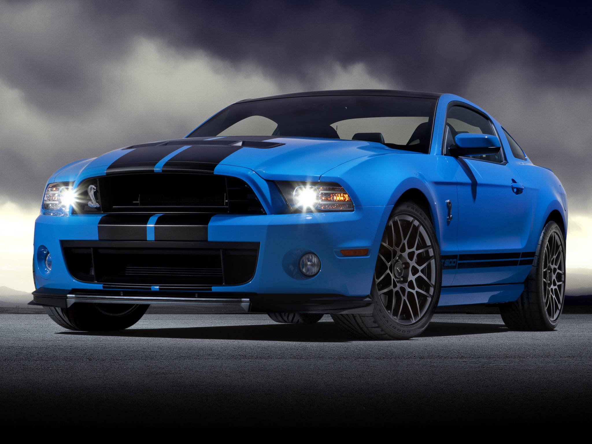 shelby iPhone 桌布 壁紙 — 一輛充滿活力的藍色福特野馬 Shelby GT500，帶有黑色賽車條紋，在戲劇性的暴風雲下亮著頭燈蓄勢待發。