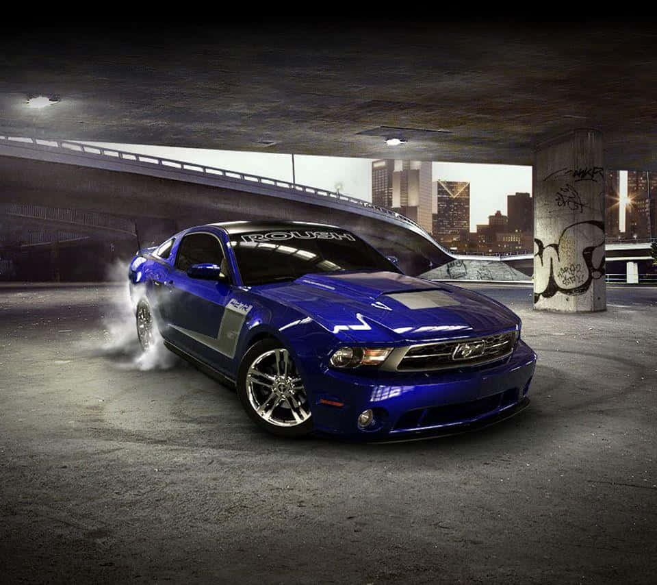 Ford Mustang Roush 桌布 壁紙 — 一輛充滿活力的藍色福特野馬在混凝土天橋下飄移，後輪冒出滾滾濃煙，背景是粗糙的城市景觀。