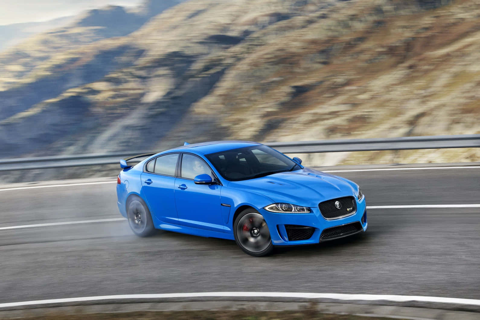 Jaguar Xfr 桌布 壁紙 — 一輛充滿活力的藍色積架跑車在蜿蜒的山路上疾馳，背景是模糊的岩石地形。