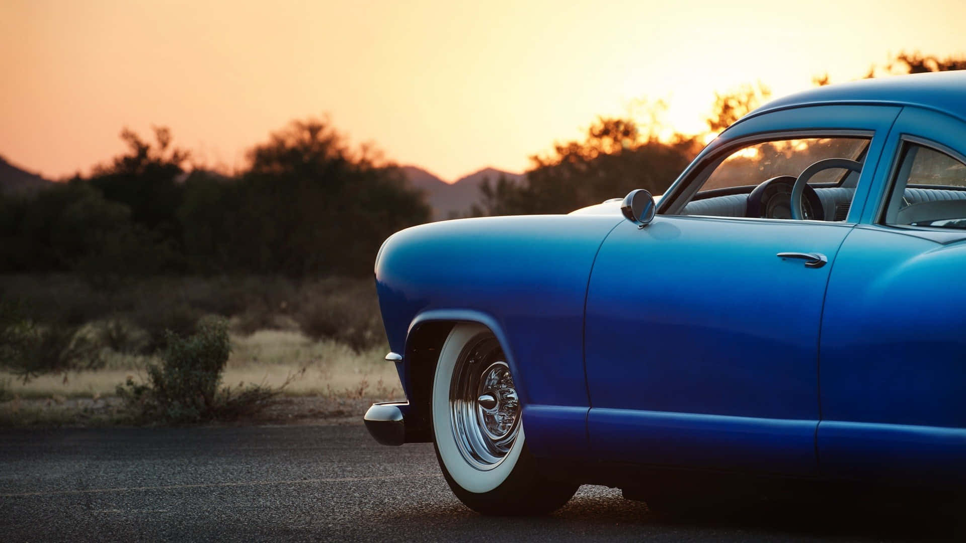 Blue Car 桌布 壁紙 — 一輛充滿活力的藍色經典汽車停在沙漠公路上，夕陽西下，溫暖的橙色天空和輪廓分明的山丘。
