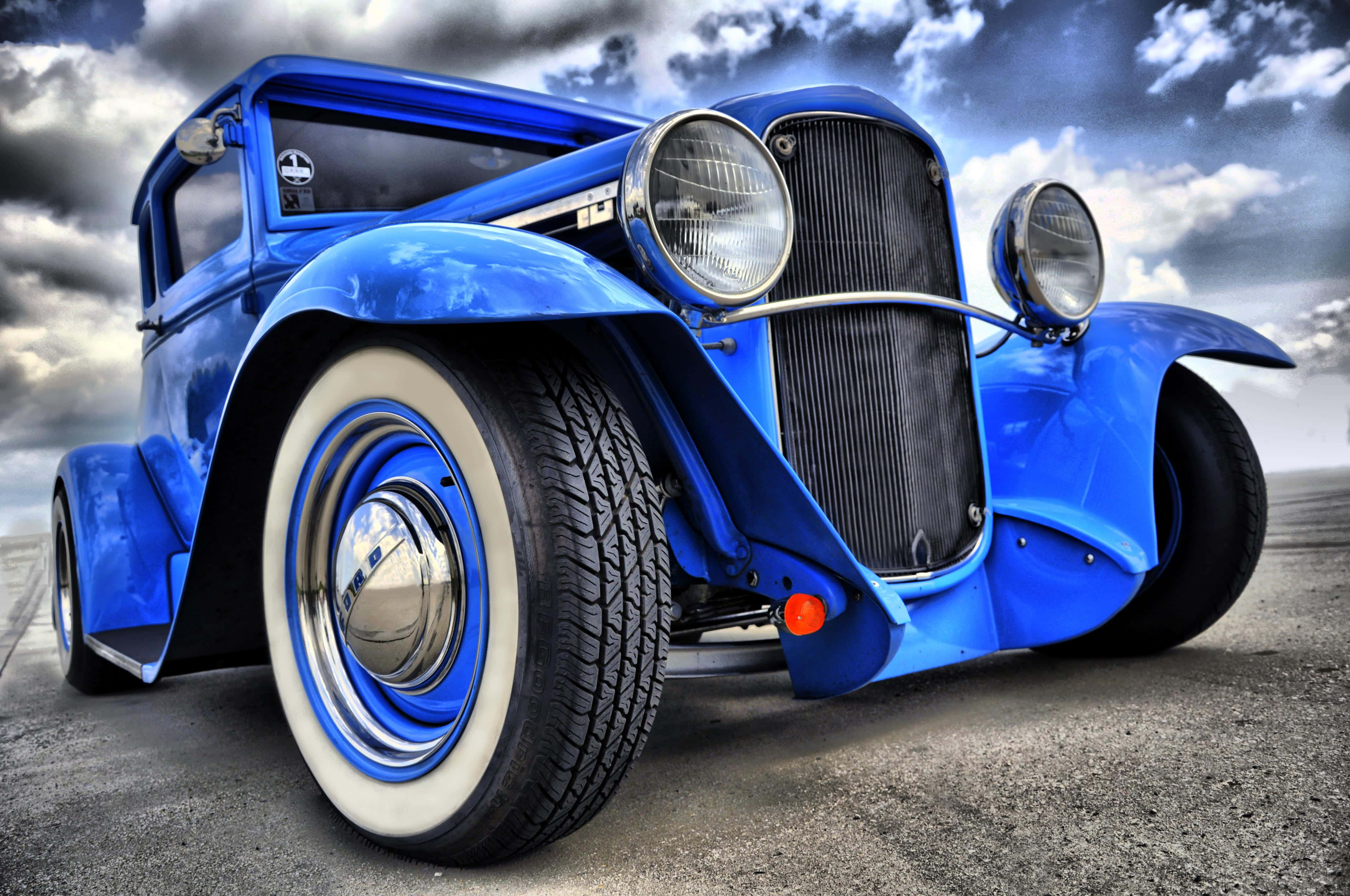 Hot Rod 桌布 壁紙 — 一輛充滿活力的藍色經典熱棒車，配備白牆輪胎和鍍鉻裝飾，在戲劇性的多雲天空下大膽矗立。