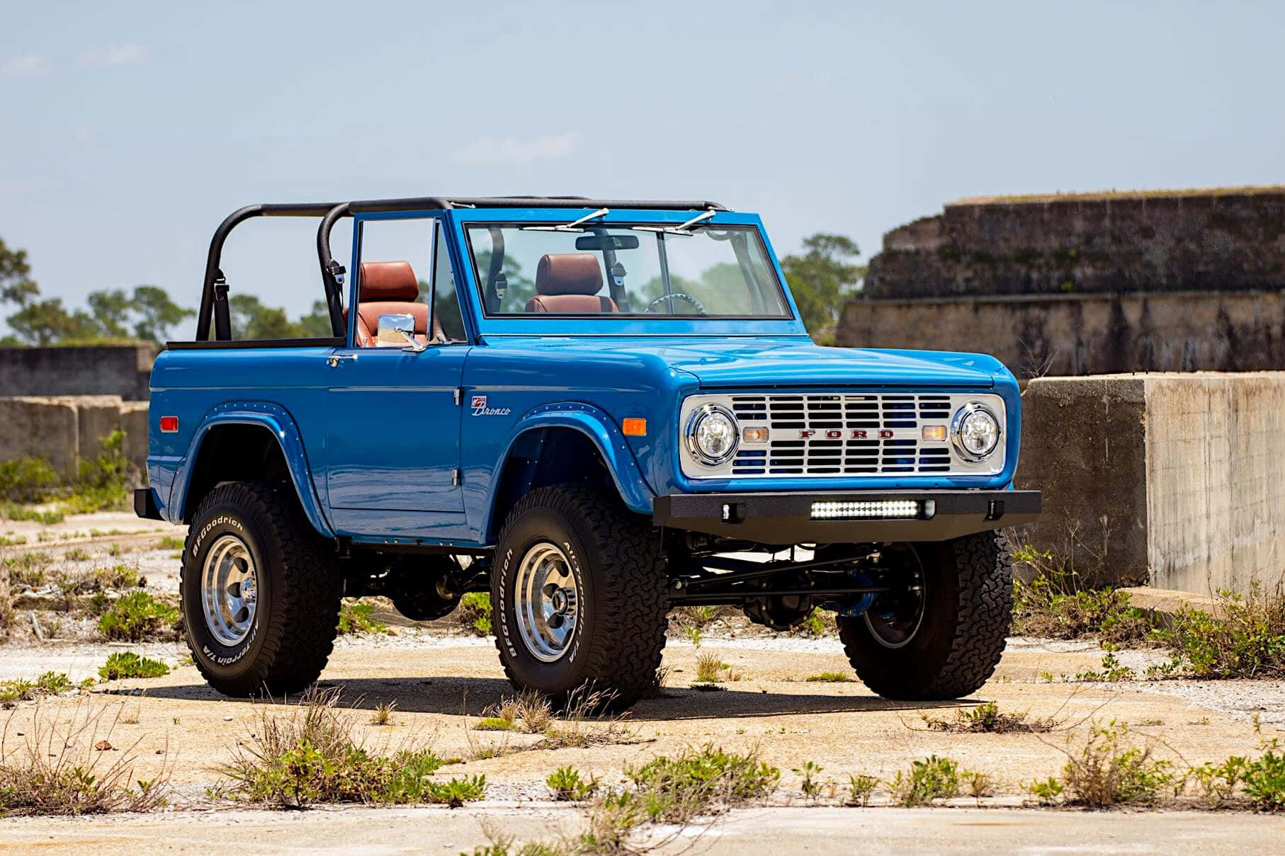 福特野馬越野車 桌布 壁紙 — 一輛充滿活力的藍色經典福特Bronco，配橙色座椅，驕傲地矗立在陽光普照的混凝土廢墟上，周圍是乾草和遠處的樹木。