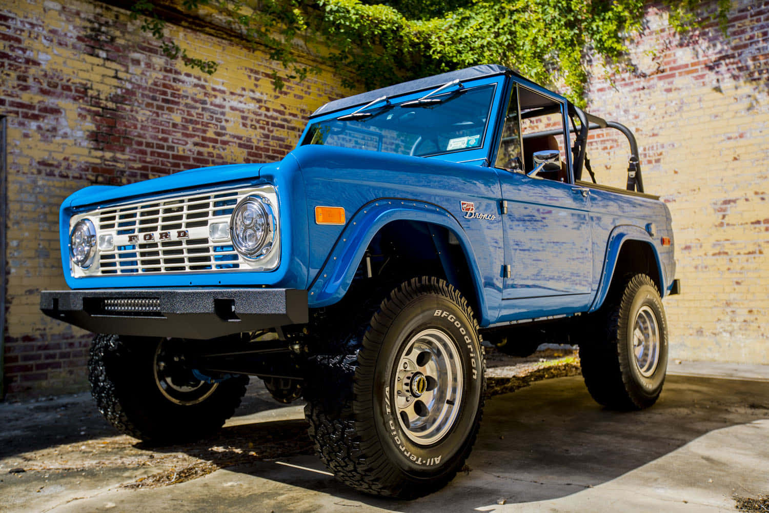 福特野馬越野車 桌布 壁紙 — 一輛充滿活力的藍色經典福特Bronco，配備超大輪胎，在明亮的陽光下自豪地停在風化的磚牆前。
