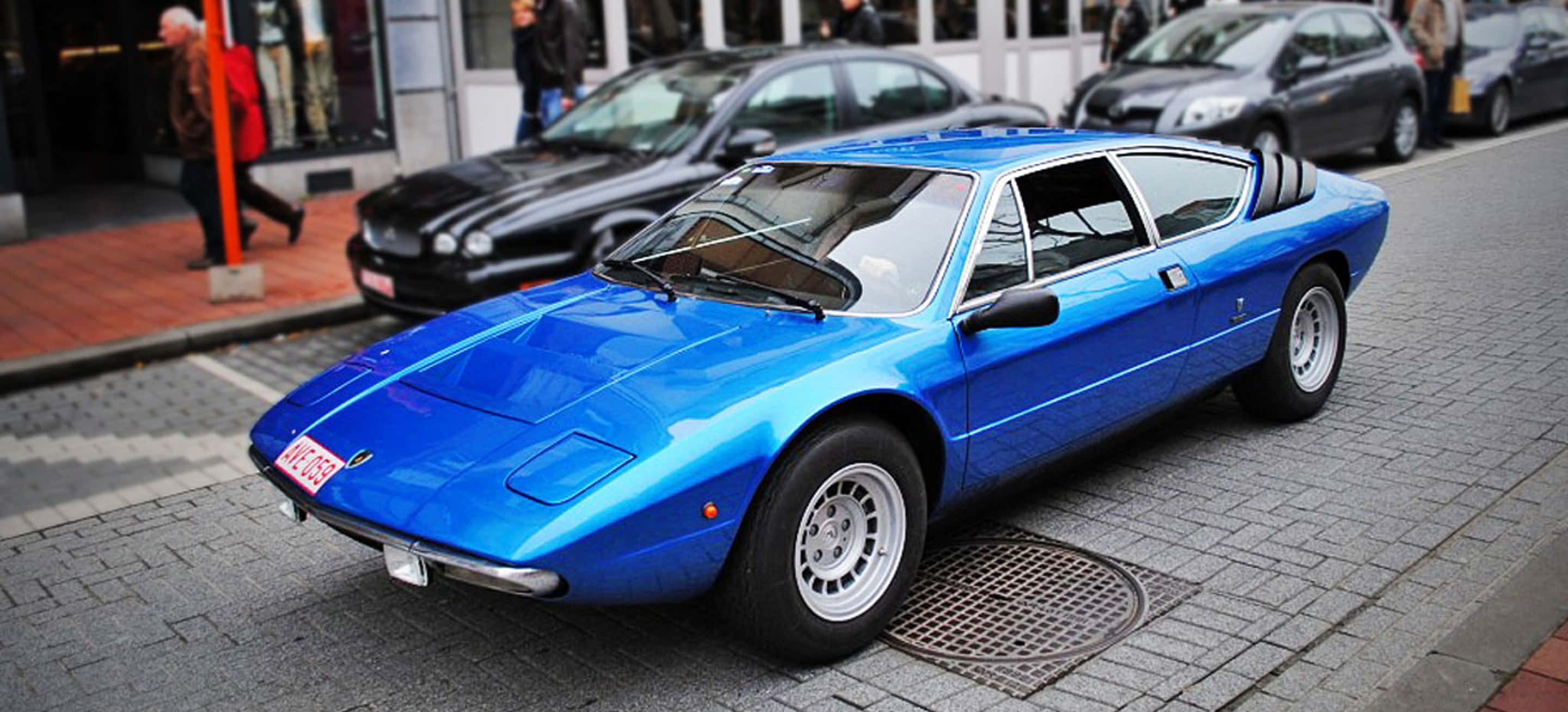 Lamborghini Urraco 桌布 壁紙 — 一輛充滿活力的藍色經典跑車停泊在鵝卵石街道上，背景有行人和現代汽車。