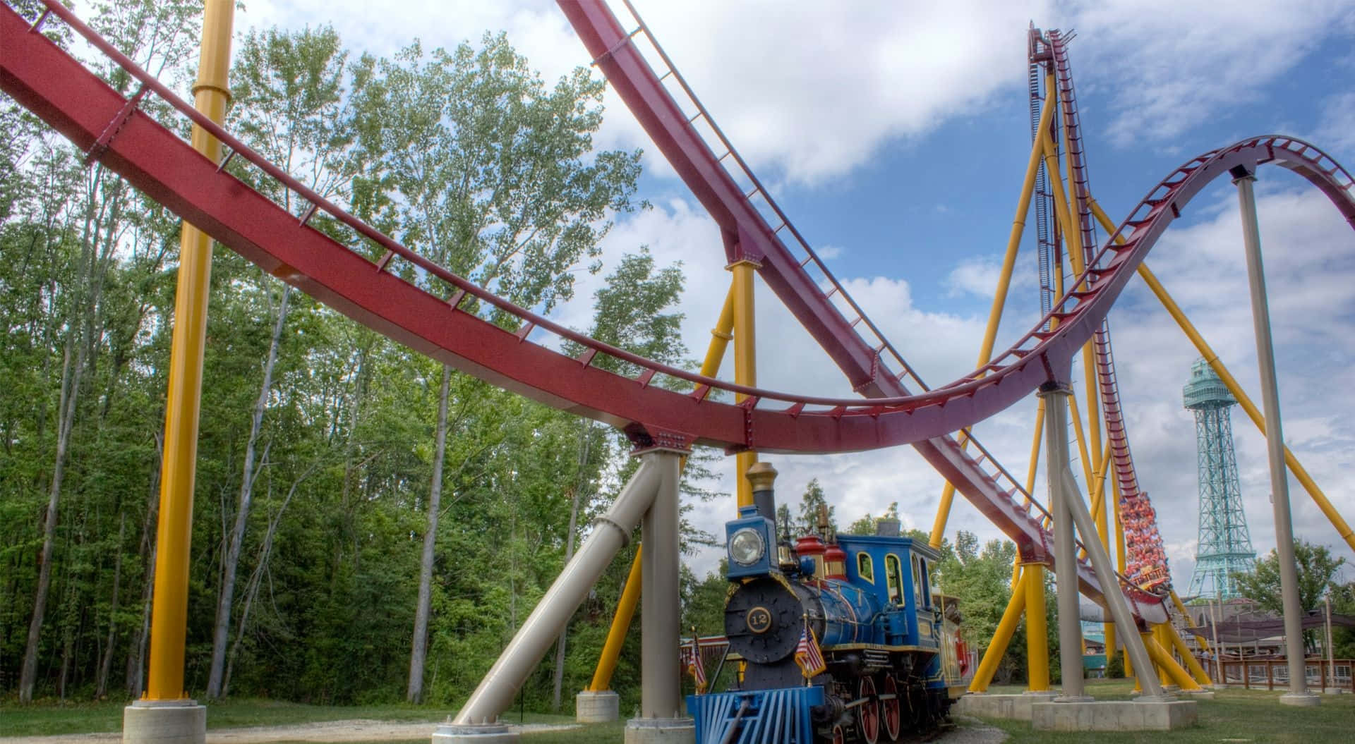 Roller Coaster 桌布 壁紙 — 一輛充滿活力的藍色蒸汽火車停在高聳的紅色和黃色雲霄飛車軌道下，背景是明亮的藍天和蓬鬆的白雲。