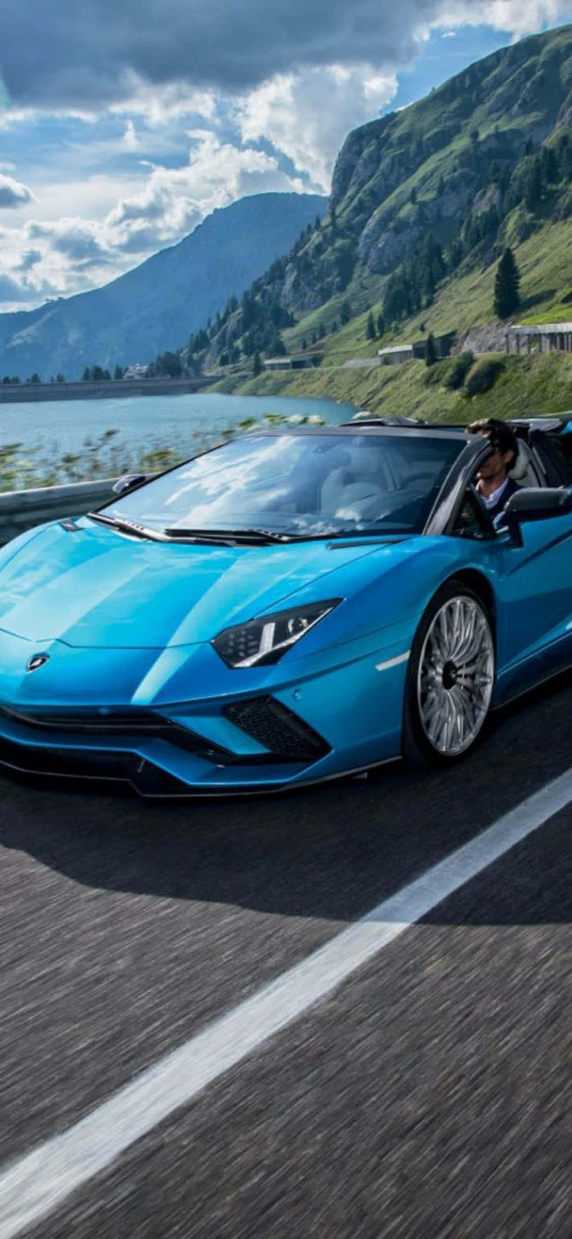 iPhone X Lamborghini 桌布 壁紙 — 一輛充滿活力的藍色藍寶堅尼 Aventador Roadster 在湖邊的山路上奔馳，天空多雲。