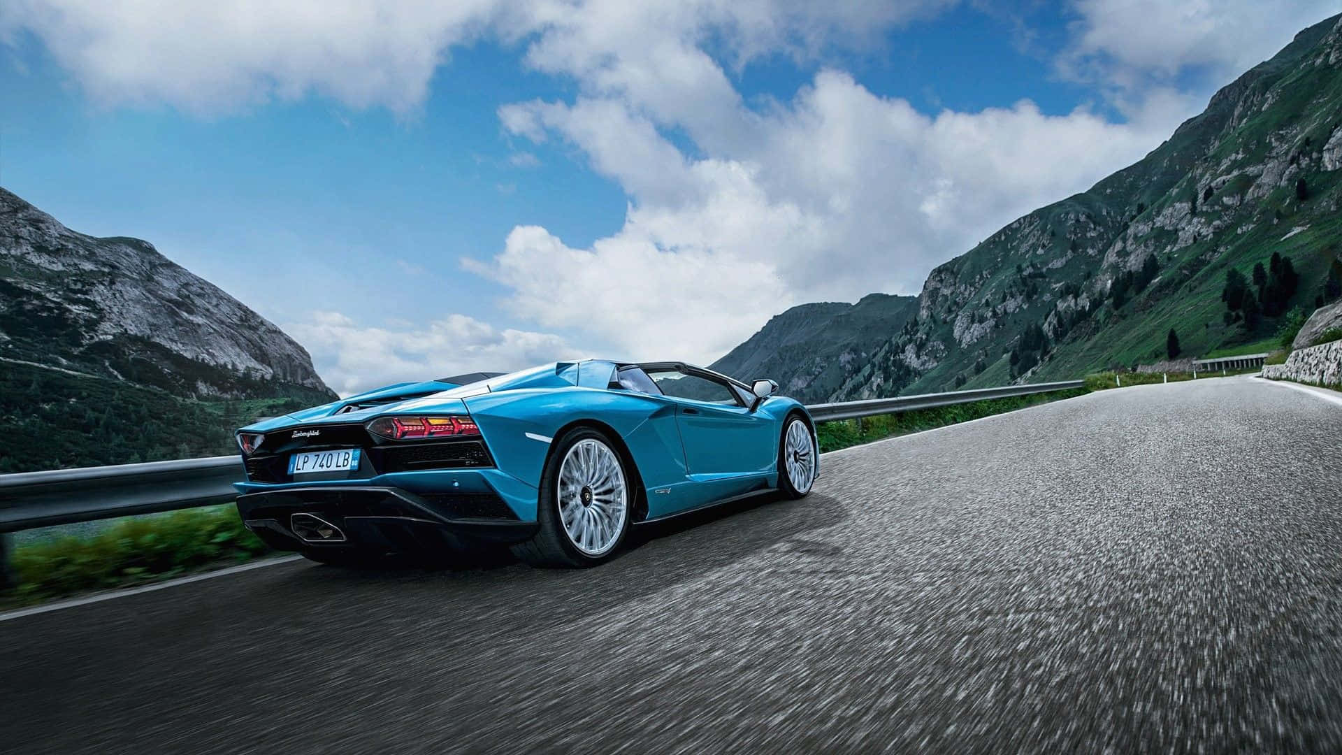 跑車 桌布 壁紙 — 一輛充滿活力的藍色藍寶堅尼 Aventador Roadster 在晴朗藍天和 fluffy 雲朵下，沿著蜿蜒的山路飛馳。