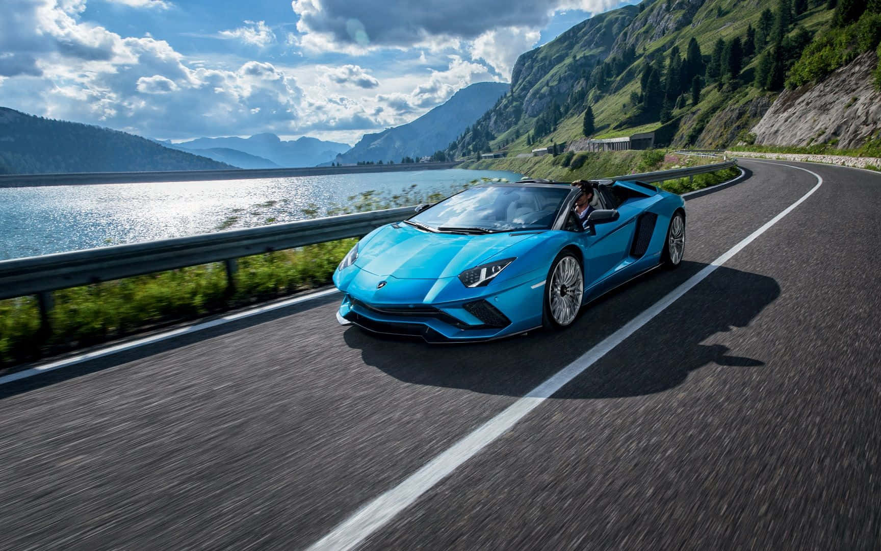 跑車 桌布 壁紙 — 一輛充滿活力的藍色藍寶堅尼 Aventador Roadster 在部分多雲的天空下，沿著蜿蜒的山路在閃閃發光的湖邊飛馳。