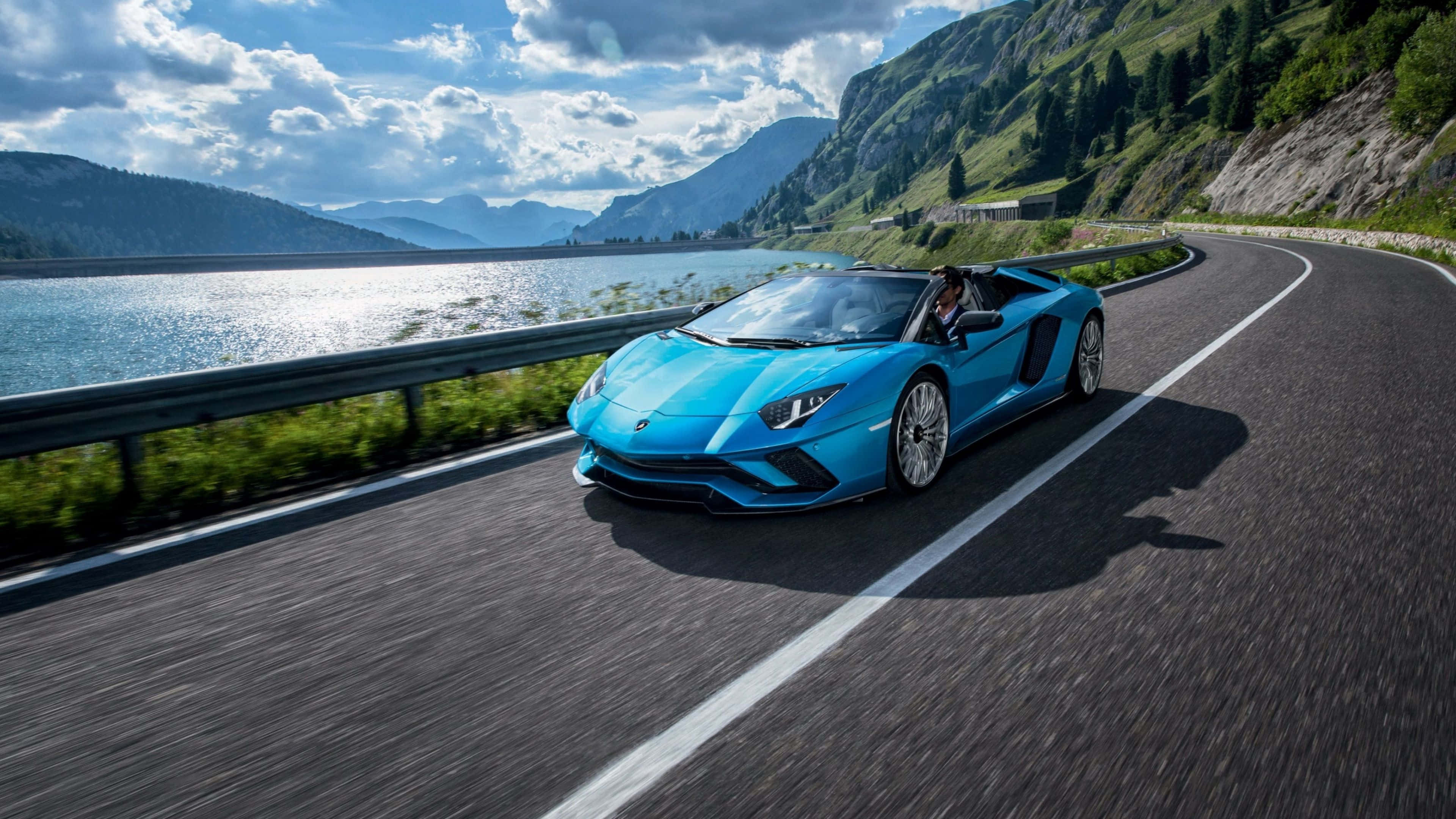 4K 藍寶堅尼 桌布 壁紙 — 一輛充滿活力的藍色藍寶堅尼 Aventador Roadster 沿著蜿蜒的山路，在波光粼粼的湖邊奔馳，天空部分多雲。