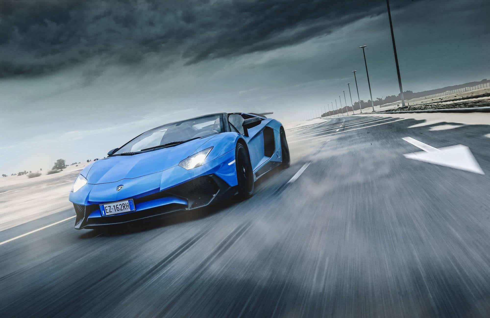 跑車 桌布 壁紙 — 一輛充滿活力的藍色藍寶堅尼 Aventador 在暴風雨的天空下，在潮濕的道路上帶著動態模糊效果高速行駛。