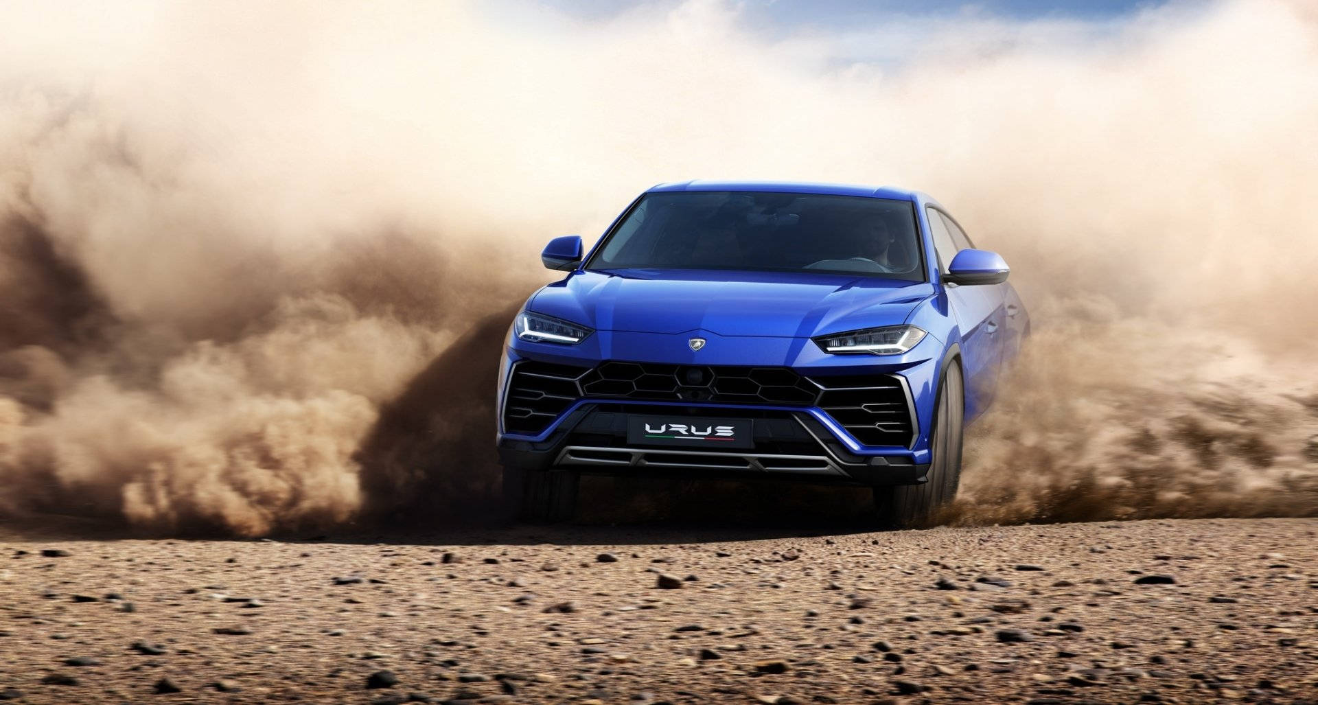 lambo 桌布 壁紙 — 一輛充滿活力的藍色藍寶堅尼 Urus 在塵土飛揚的沙漠地形中咆哮，揚起巨大的沙塵雲。