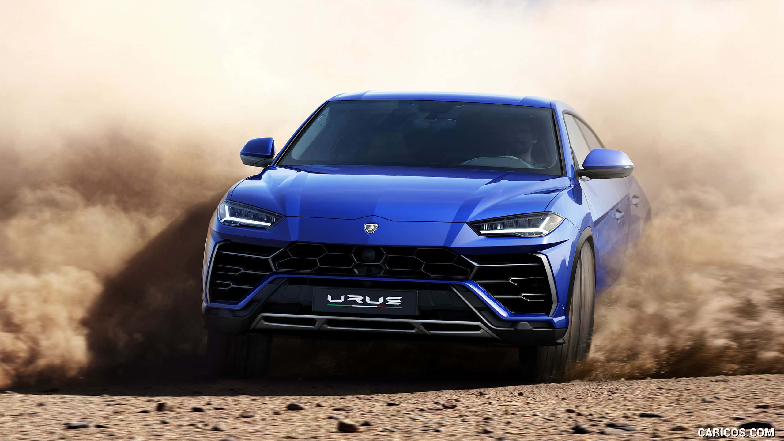 Lamborghini Urus 桌布 壁紙 — 一輛充滿活力的藍色藍寶堅尼 Urus SUV 在崎嶇的沙漠地形上向前衝刺，揚起塵土。