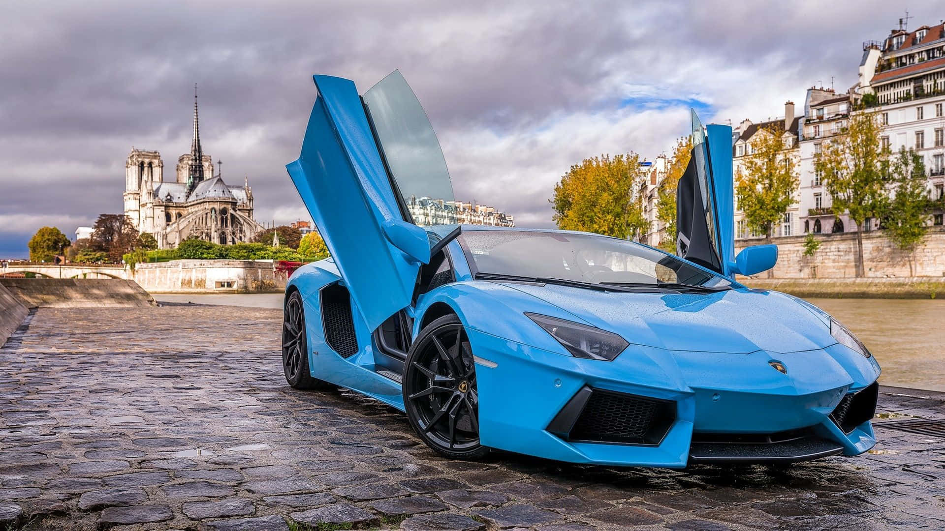 跑車 桌布 壁紙 — 一輛充滿活力的藍色藍寶堅尼Aventador，剪刀門打開，停在巴黎聖母院前的鵝卵石河岸上，天空多雲。