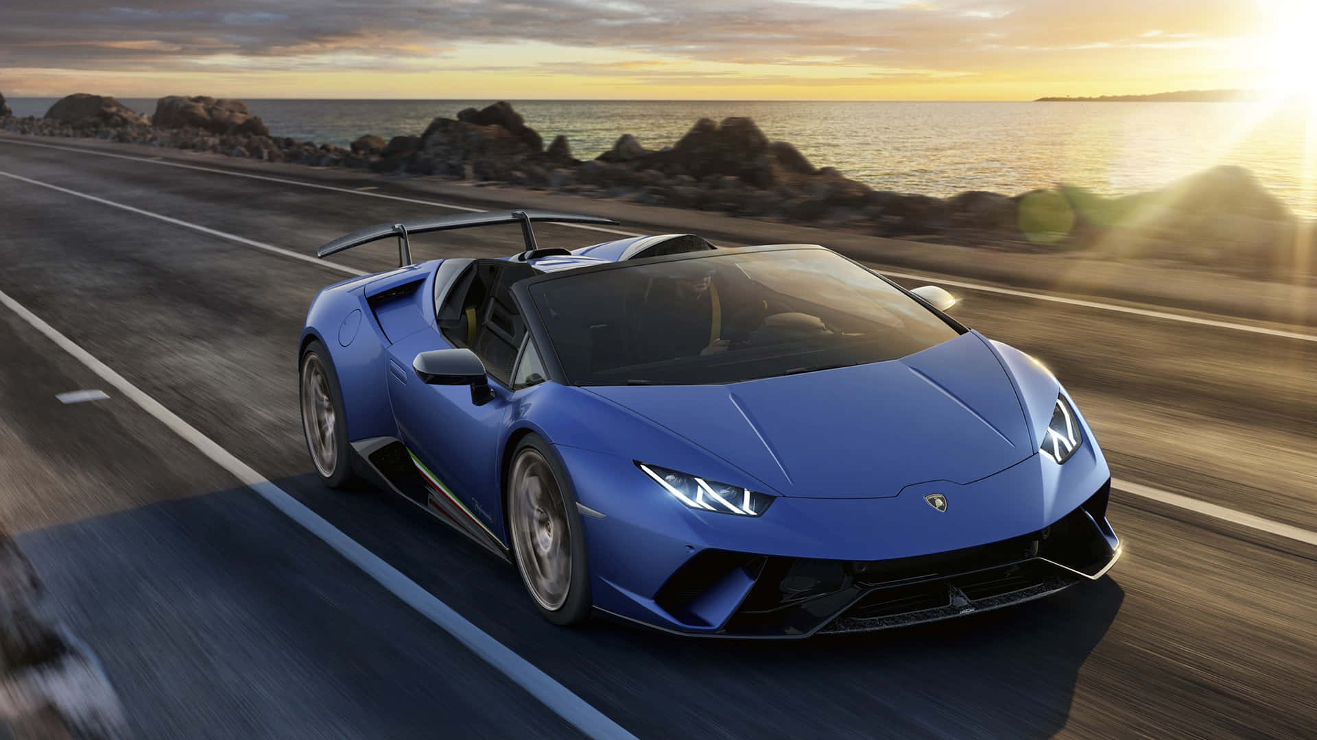 Blue Car 桌布 壁紙 — 一輛充滿活力的藍色藍寶堅尼Huracán Spyder在日落時分沿著海岸公路疾駛，背景是海洋和岩石。