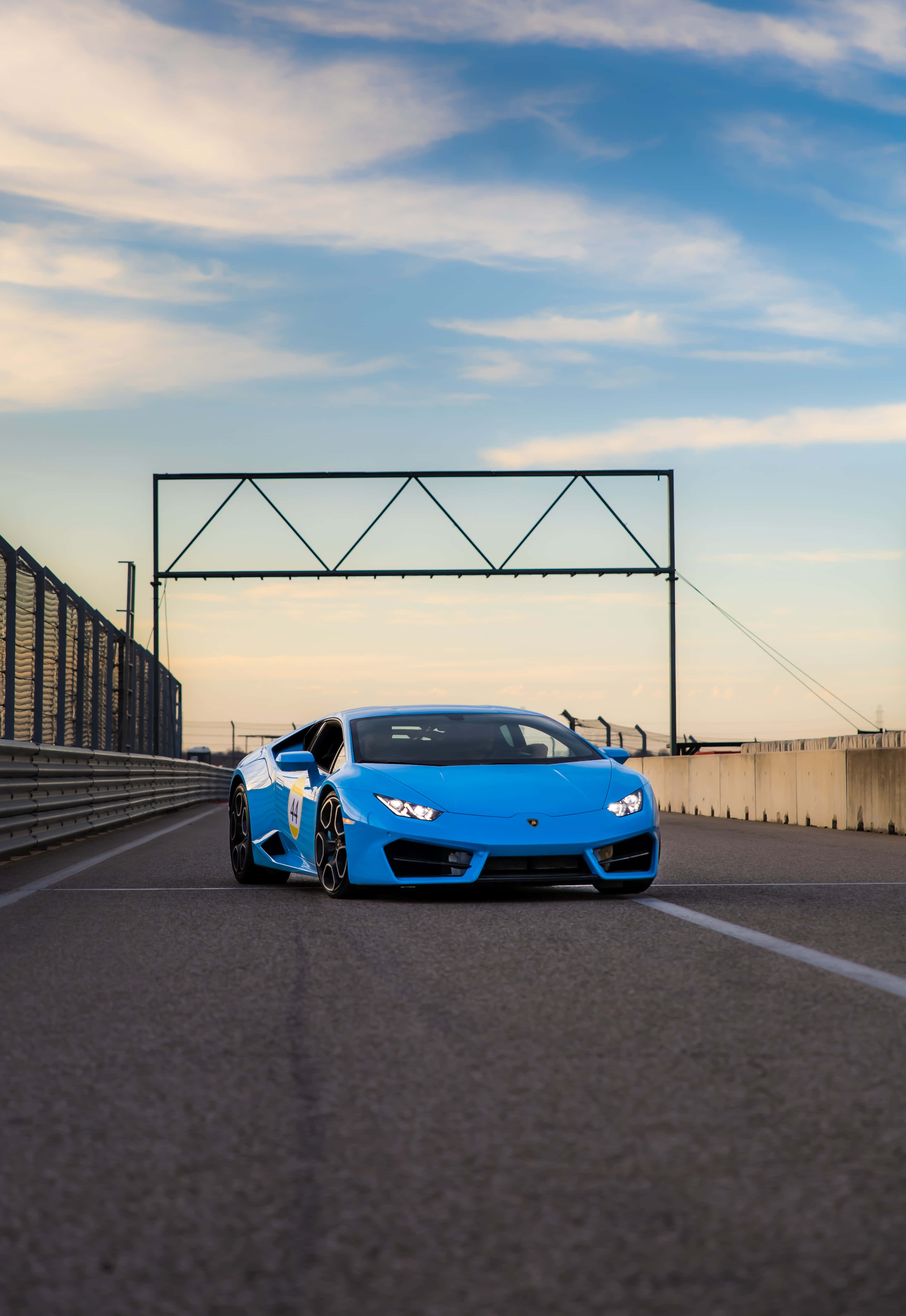Blue Lamborghini iPhone 桌布 壁紙 — 一輛充滿活力的藍色藍寶堅尼跑車在柔和的金色日落天空下沿著賽道飛馳。