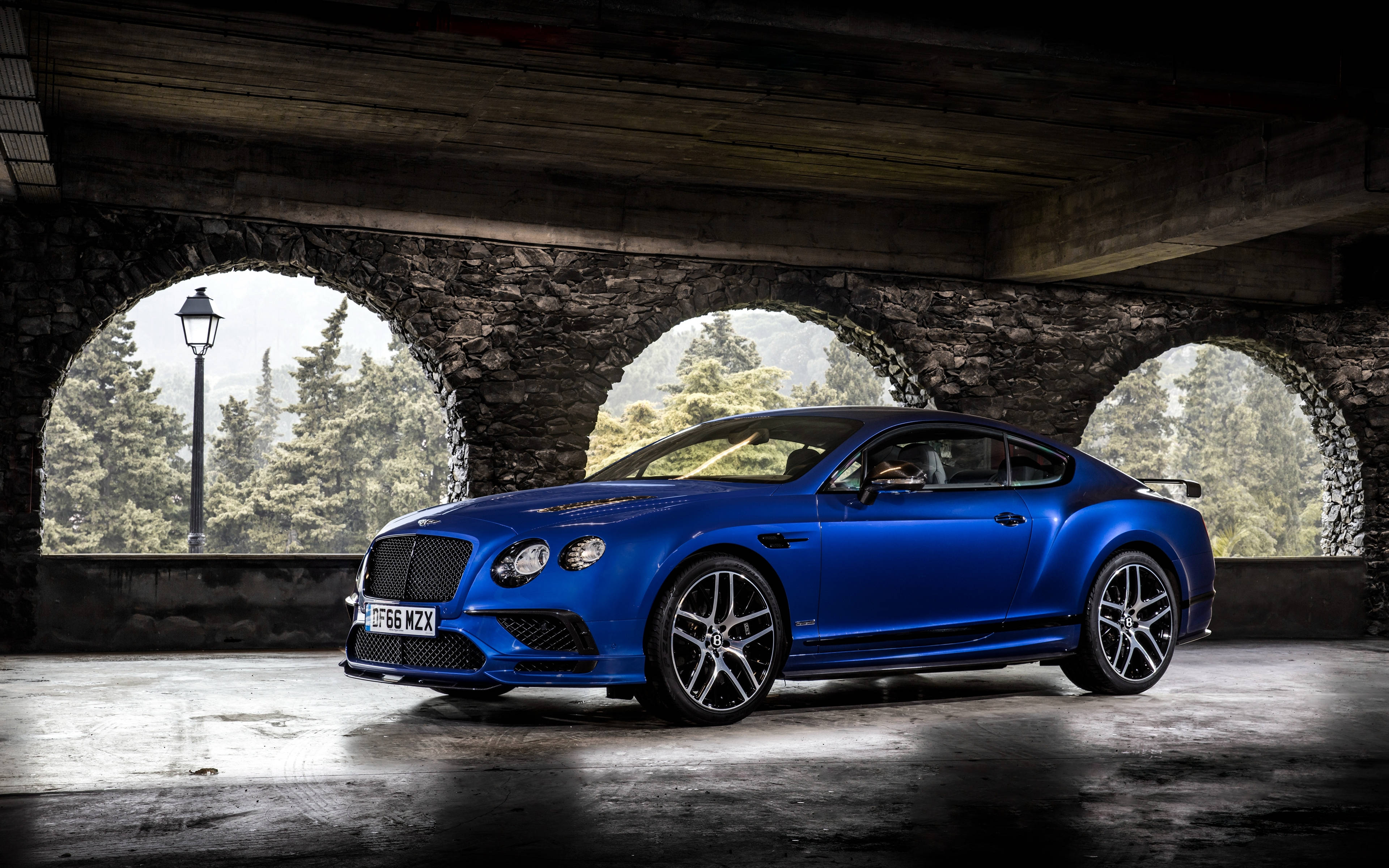 bentley HD 桌布 壁紙 — 一輛充滿活力的藍色賓利 Continental GT Speed 在石拱門下，在戲劇性的燈光下閃閃發光，可欣賞森林景色。
