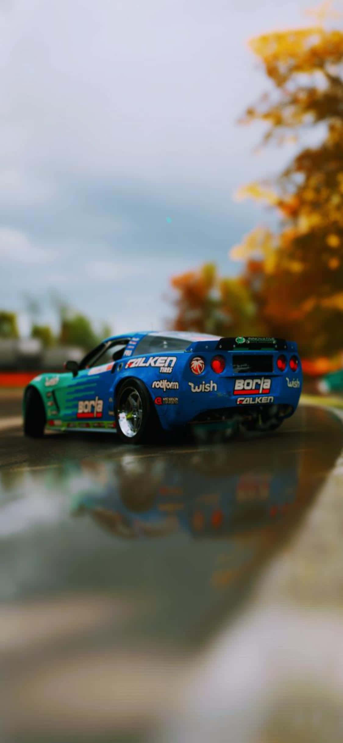 iPhone Xs Forza Horizon 4 桌布 壁紙 — 一輛充滿活力的藍色賽車帶有贊助商貼紙，在多雲的天空下沿著濕滑的賽道疾馳，賽道反射出秋天的樹木。