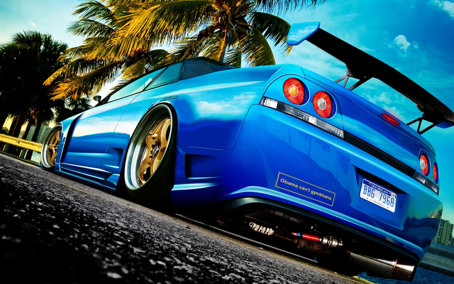 R33 GT-R 桌布 壁紙 — 一輛充滿活力的藍色跑車，配備金色輪圈和大型後尾翼，在陽光明媚的日子停放在棕櫚樹下。
