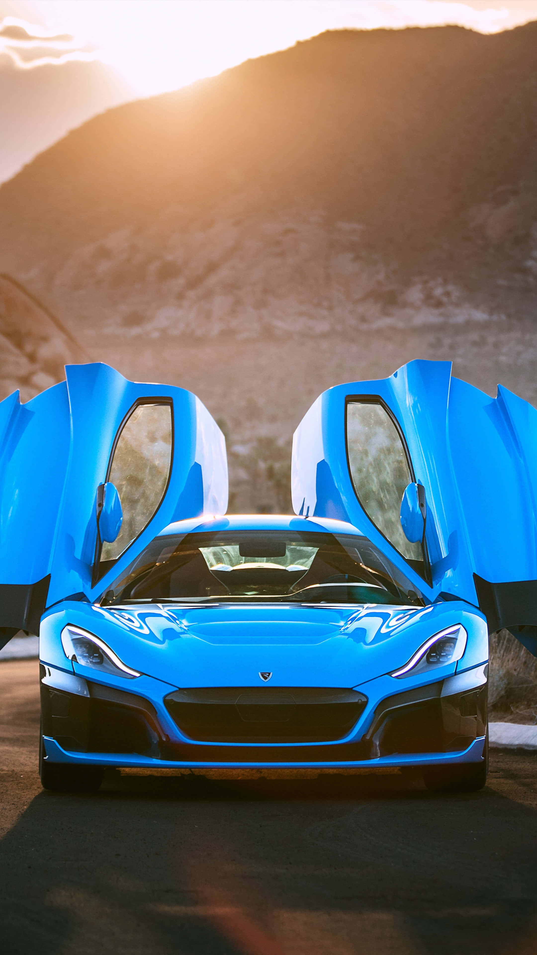 Rimac 桌布 壁紙 — 一輛充滿活力的藍色跑車，鷗翼車門敞開，在黃金時段停泊在沙漠公路上，背景是山脈。