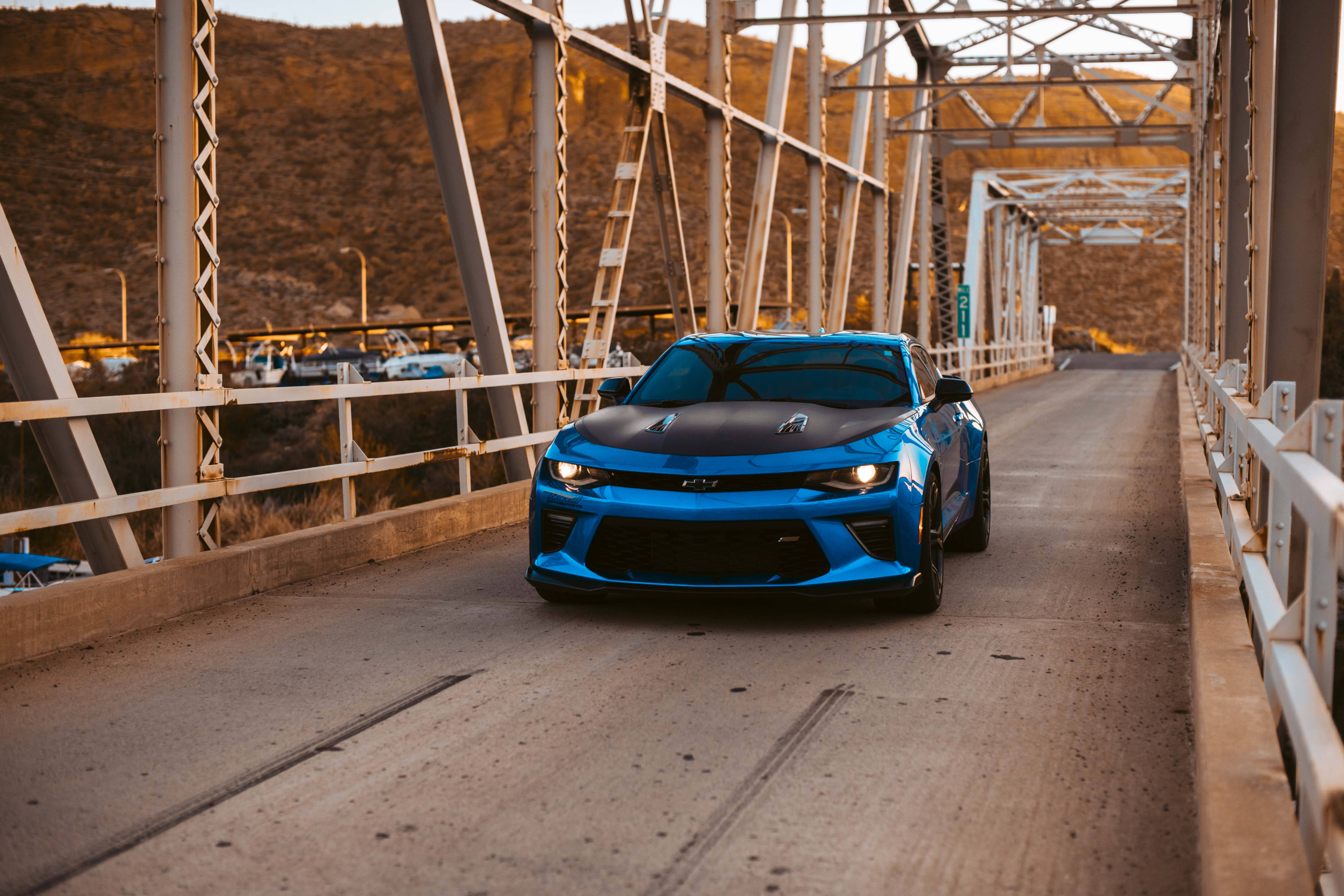 Chevrolet 桌布 壁紙 — 一輛充滿活力的藍色跑車，黑色引擎蓋，在黃金時段駛向觀眾，行駛在金屬桁架橋上。