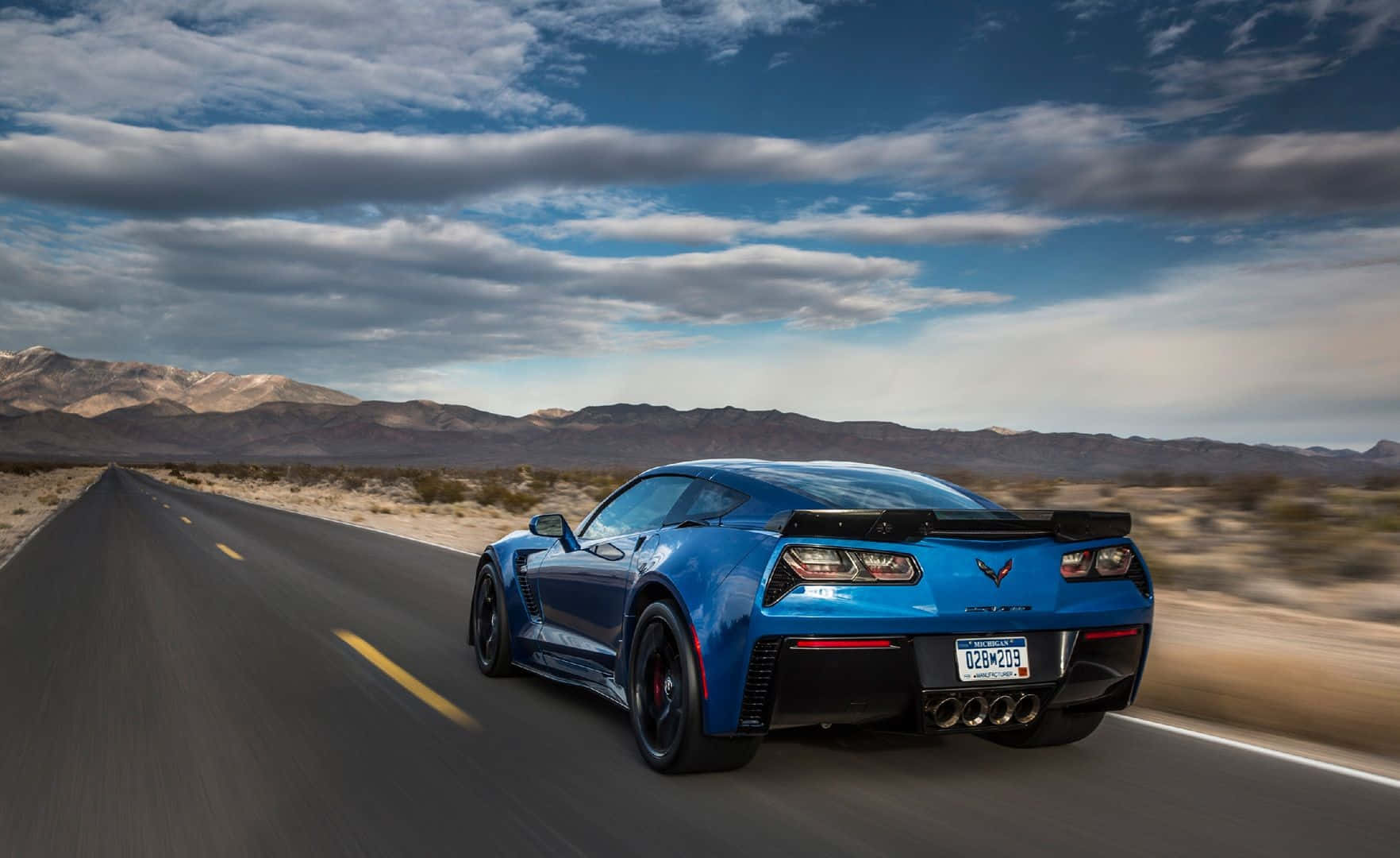 Chevrolet Corvette Z06 桌布 壁紙 — 一輛充滿活力的藍色跑車在沙漠公路上飛馳，背景是山脈和多雲的天空。