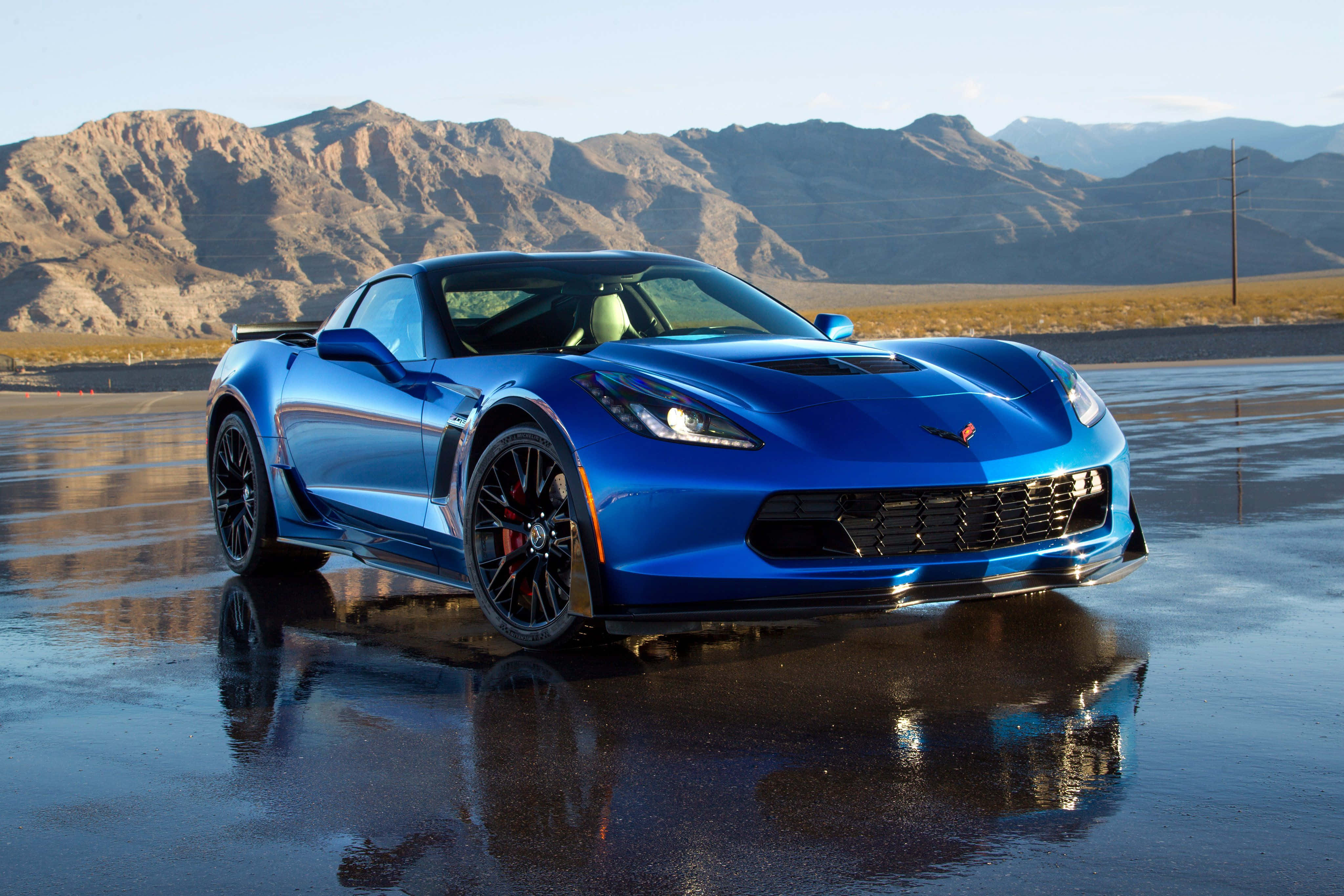 Chevrolet Corvette Z06 桌布 壁紙 — 一輛充滿活力的藍色跑車在潮濕的沙漠賽道上閃閃發光，背景是崎嶇的山脈，天空晴朗。