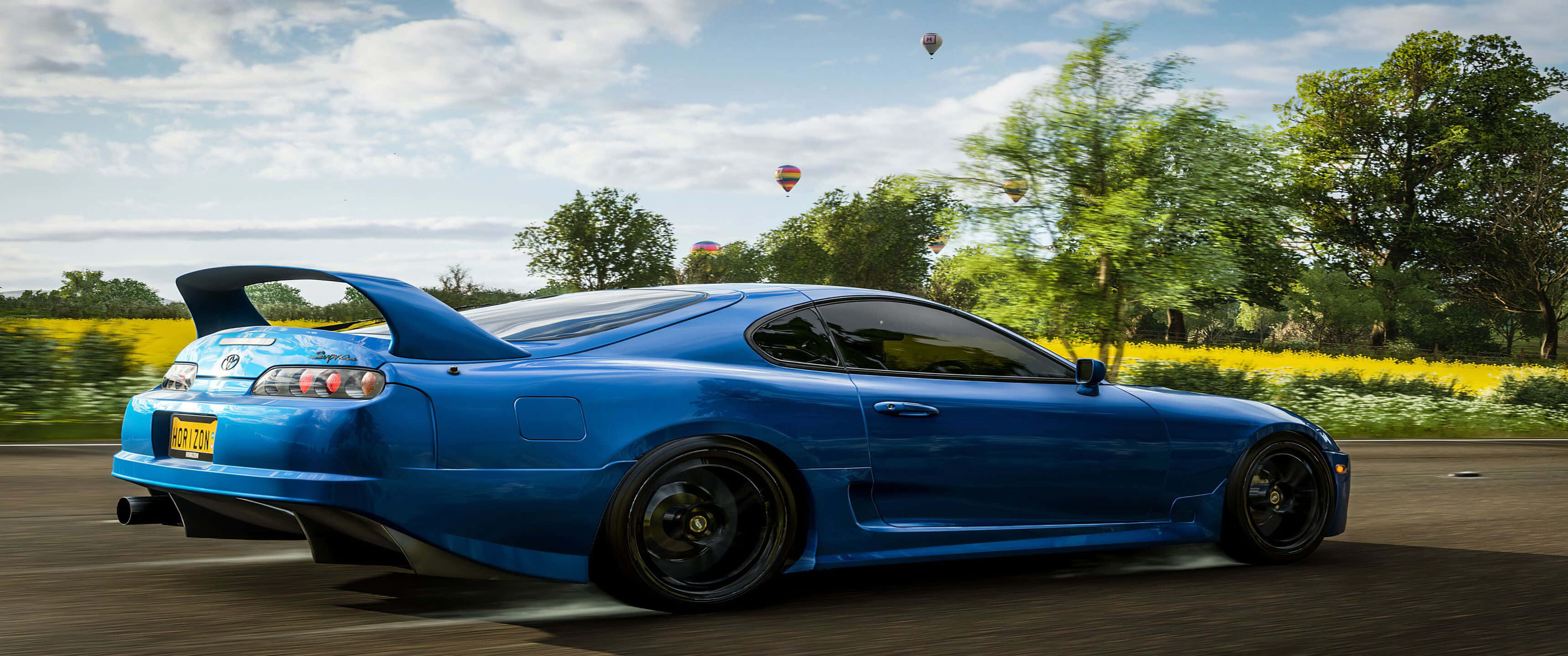 Toyota Supra 桌布 壁紙 — 一輛充滿活力的藍色跑車在部分多雲的天空下，以模糊的動態沿著道路飛馳，經過黃色的田野和樹木。