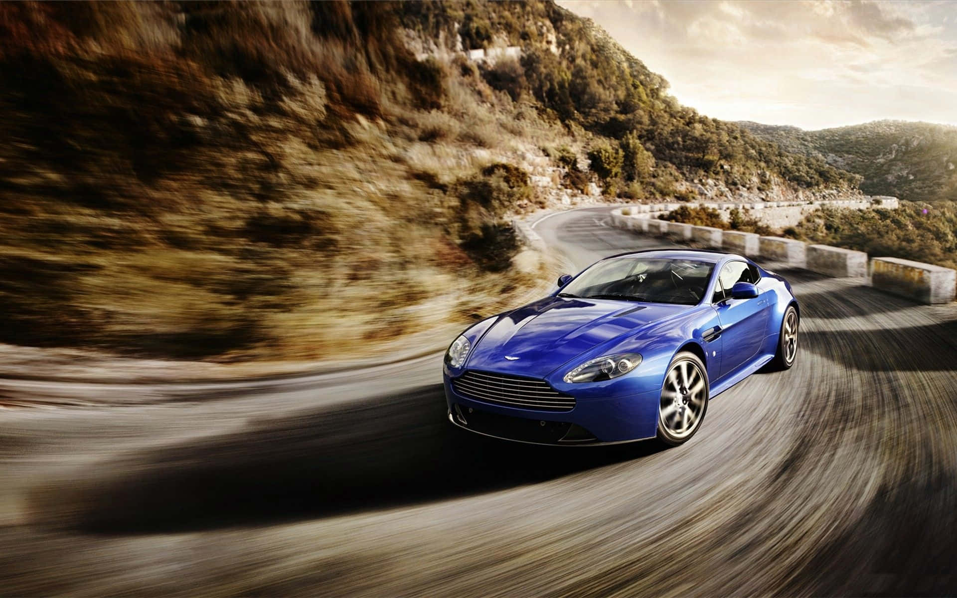 Aston Martin V12 Vantage 桌布 壁紙 — 一輛充滿活力的藍色跑車沿著蜿蜒的山路疾馳，帶有模糊的動態和金色的陽光。