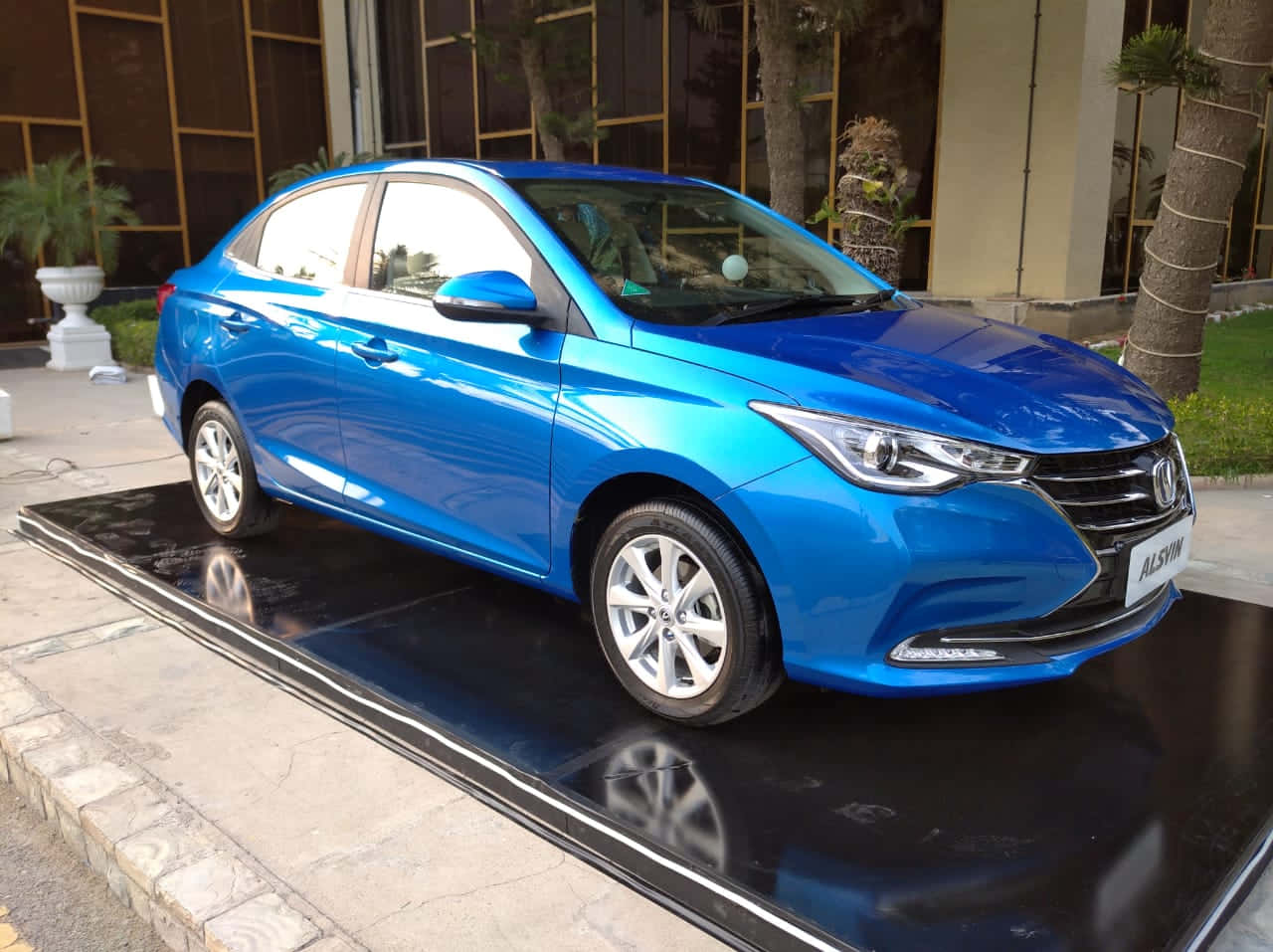 Changan 桌布 壁紙 — 一輛充滿活力的藍色轎車在日光下閃閃發光，展示在現代建築外帶有棕櫚樹的光澤黑色平台上。