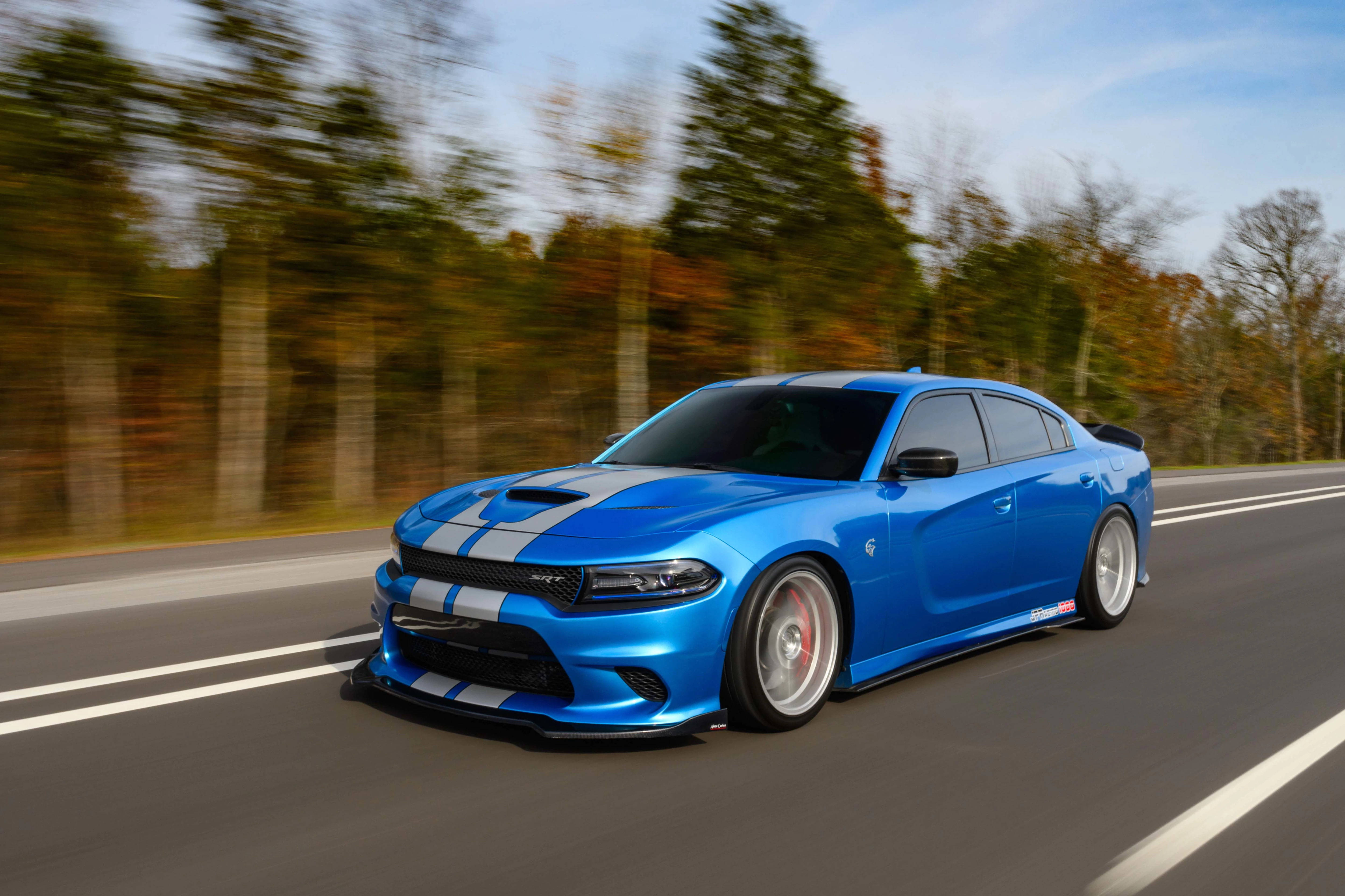 Mopar 桌布 壁紙 — 一輛充滿活力的藍色道奇Charger，帶有白色賽車條紋，在晴朗的天空下，沿著兩旁秋樹的道路飛馳。