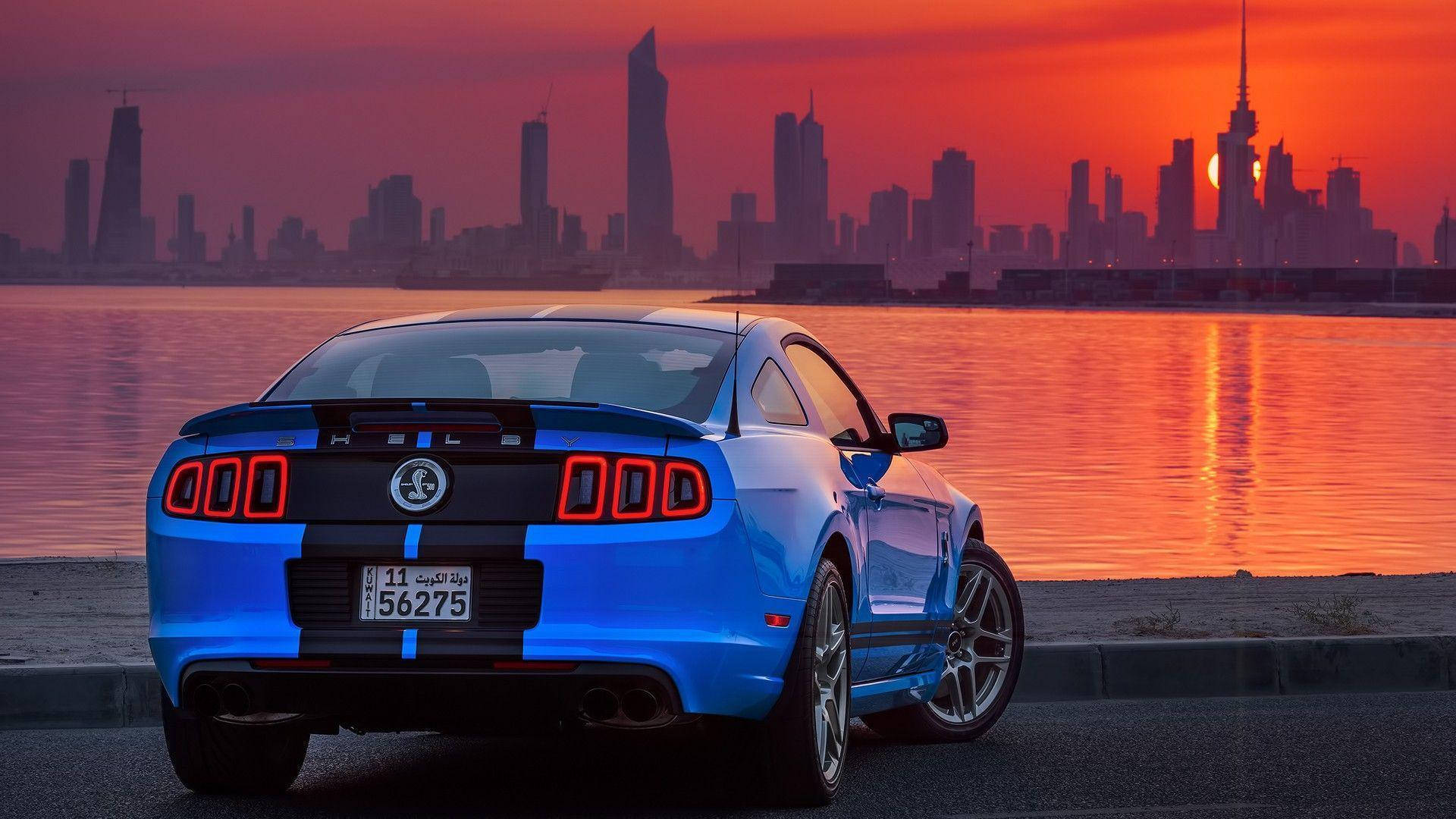 Kuwait 桌布 壁紙 — 一輛充滿活力的藍色野馬跑車在日落時停在水邊，背景是閃爍的城市天際線。