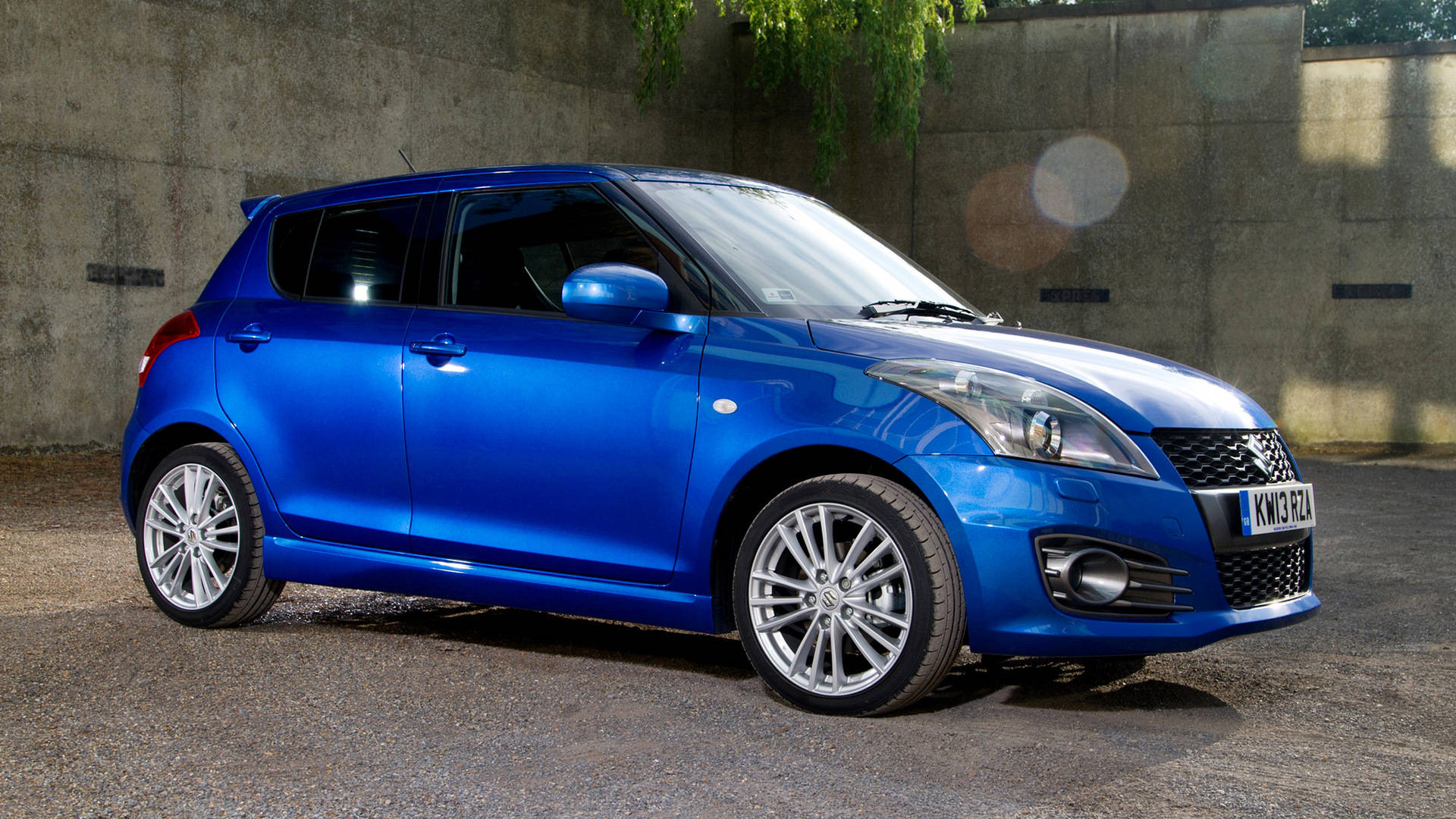Suzuki 桌布 壁紙 — 一輛充滿活力的藍色鈴木 Swift 停泊在碎石上，背景是帶紋理的混凝土牆，陽光斑駁，鏡頭眩光。