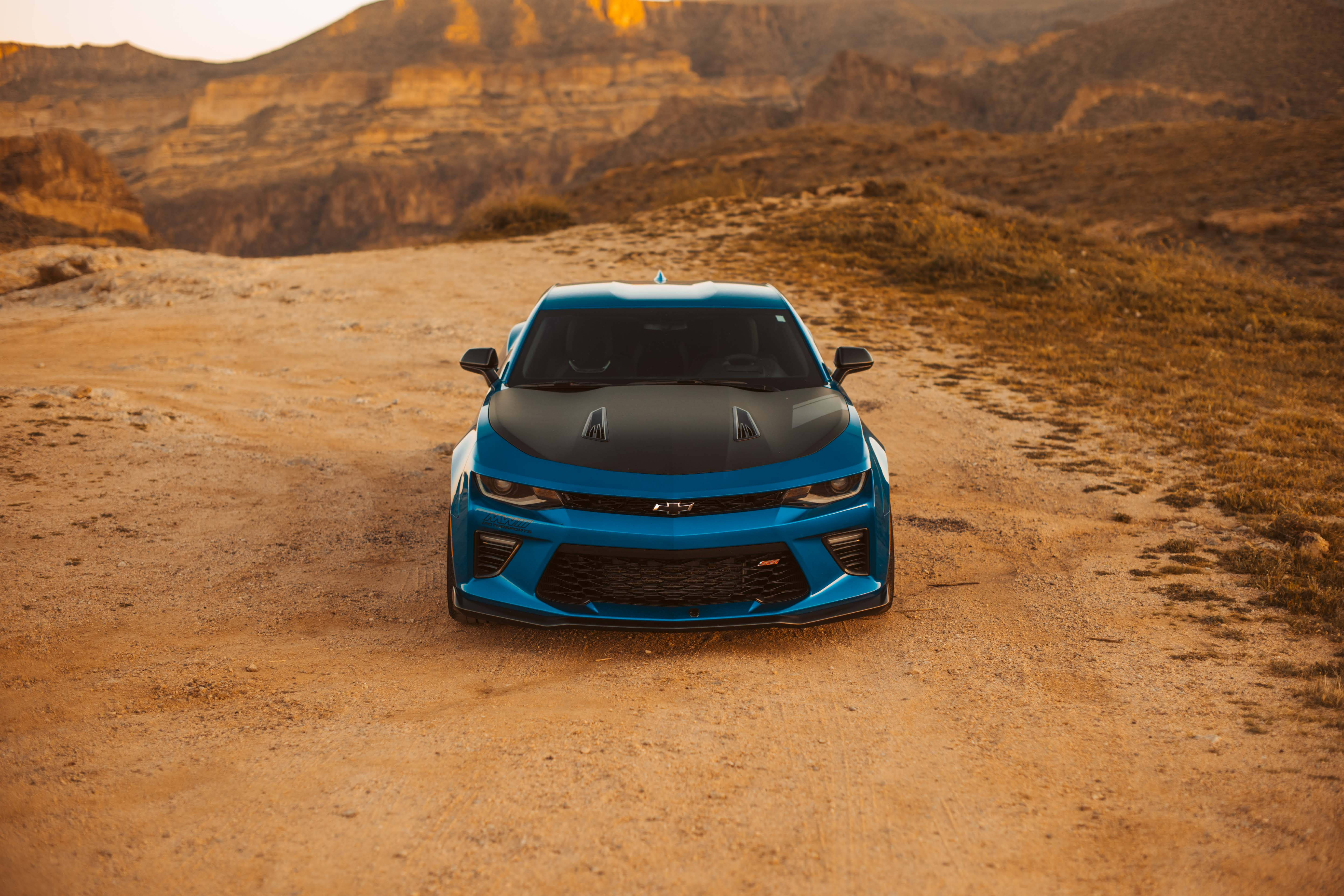 Chevrolet 桌布 壁紙 — 一輛充滿活力的藍色雪佛蘭 Camaro SS 黑色引擎蓋，停在塵土飛揚的沙漠小徑上，背景是崎嶇的峽谷懸崖，沐浴在金色的日落光中。